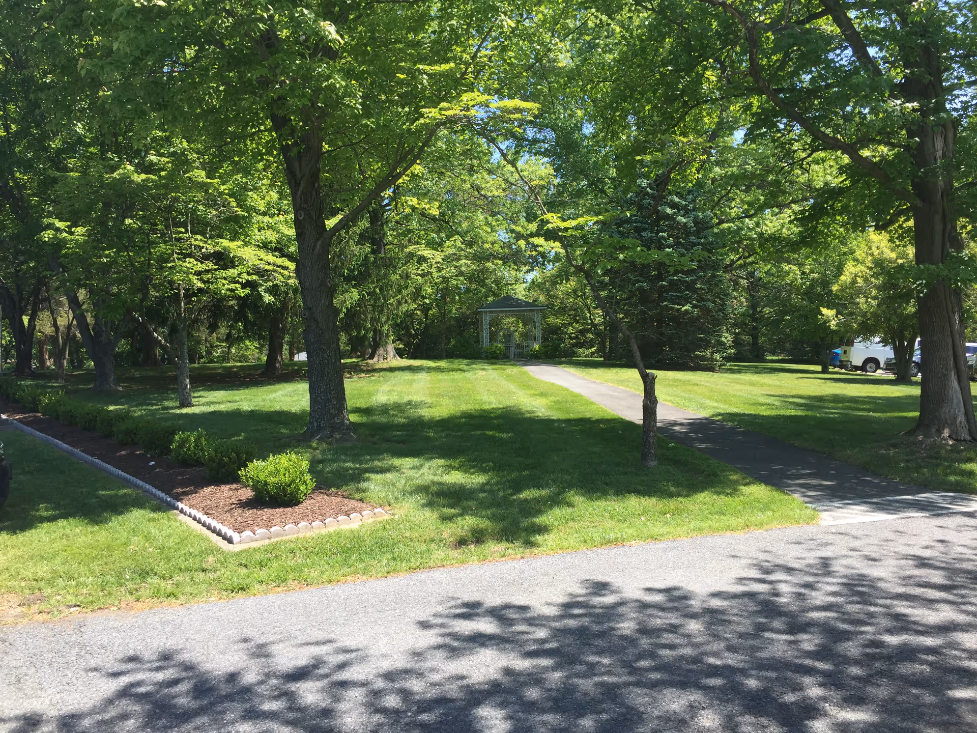 Tree-shaded front lawn with a paved walkway leading to a small gazebo and a parked vehicle at the right.