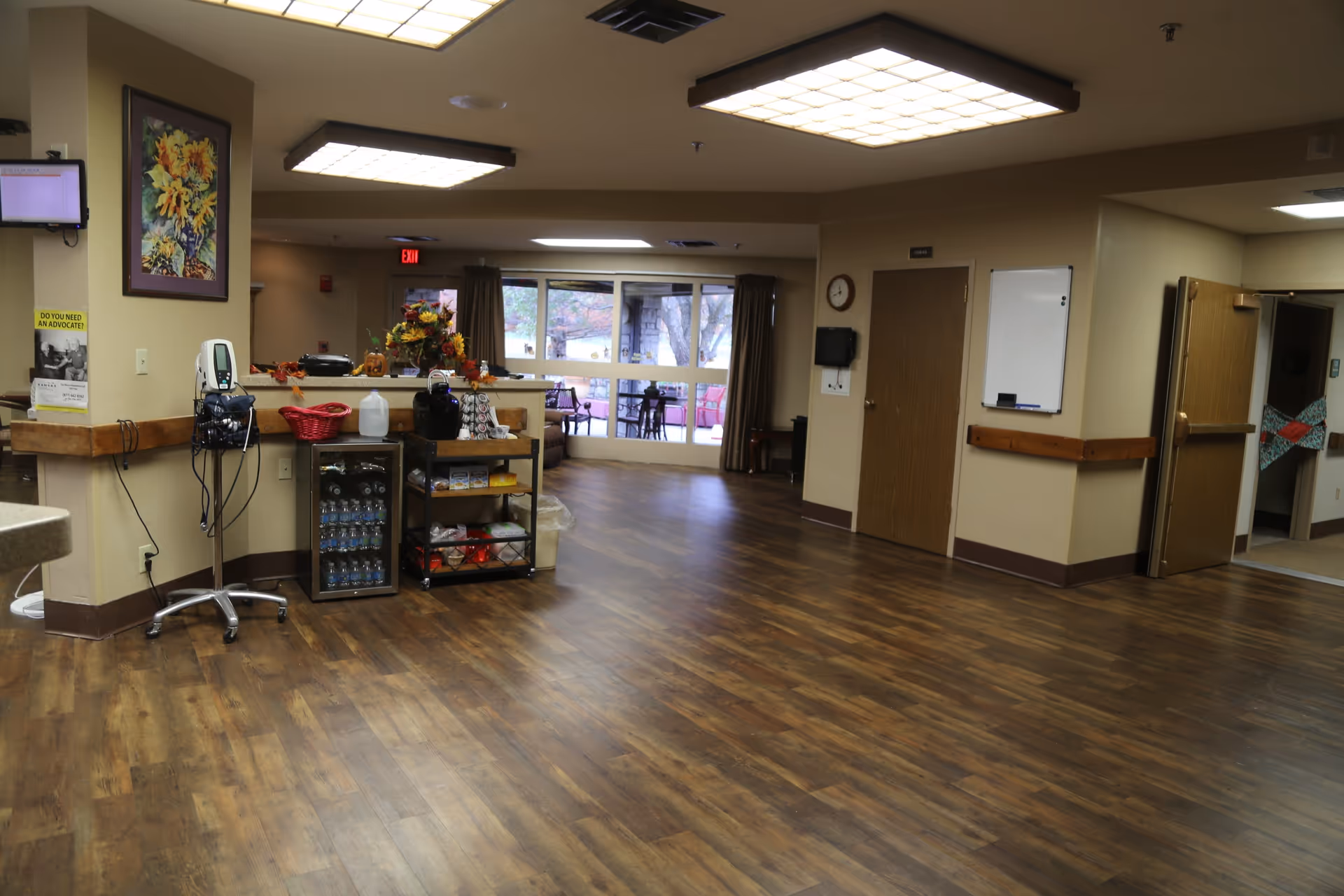 Spacious common area of a senior living facility with wood floors, a reception counter, medical equipment and seating near large windows.