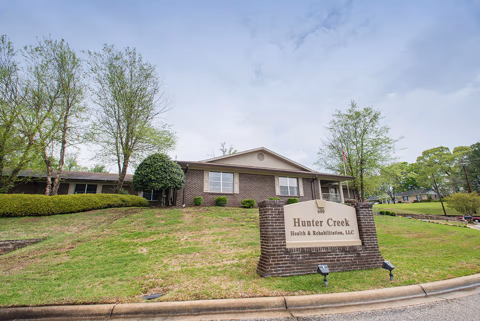 Exterior view of Hunter Creek Health and Rehabilitation, LLC building with a brick sign in front displaying the facility's name. The building is surrounded by green grass, trees, and shrubs under a partly cloudy sky.