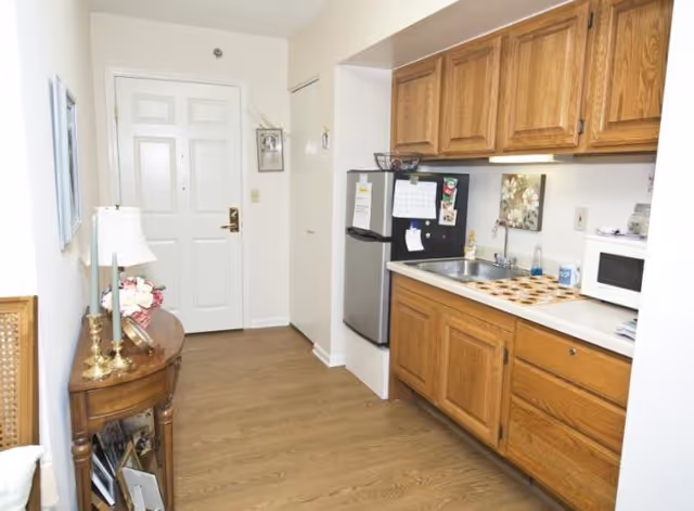 Interior view of a small kitchenette area with wooden cabinets, a stainless steel sink, a mini refrigerator, a microwave, and a countertop with a decorative mat. To the left, there is a wooden side table with a lamp, flowers, and framed photos. The floor is wooden, and a white door is visible at the end of the hallway.