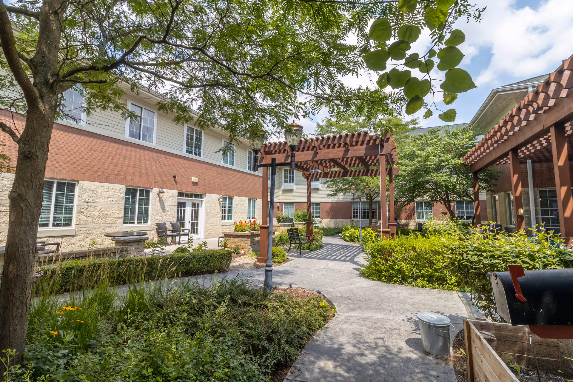 Outdoor courtyard area of a senior living facility with a paved walkway, green plants, trees, benches, and wooden pergolas providing shade. The building surrounding the courtyard has multiple windows and a mix of brick and light-colored siding.