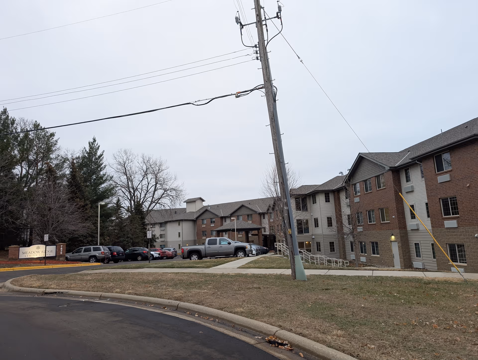 Exterior view of a three-story brick-and-siding senior living building with parked cars, a utility pole, and a Meadow Ridge sign under an overcast sky.