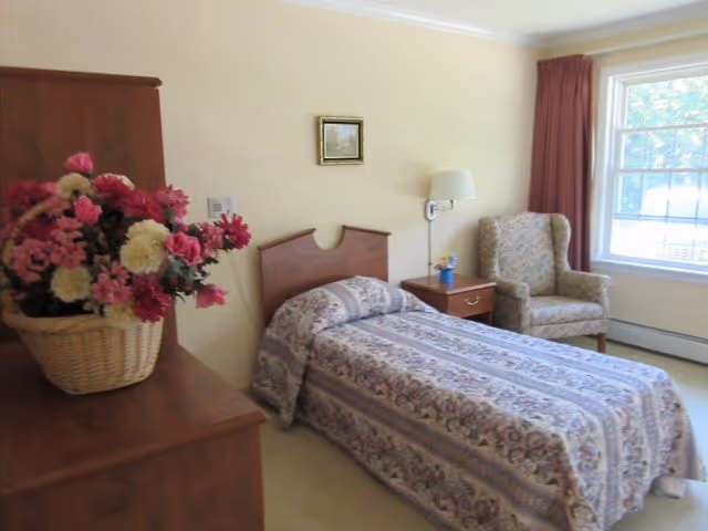 Sunlit single-occupancy bedroom with a floral-covered twin bed, wooden dresser topped with a basket of flowers, a nightstand, and an armchair by the window.