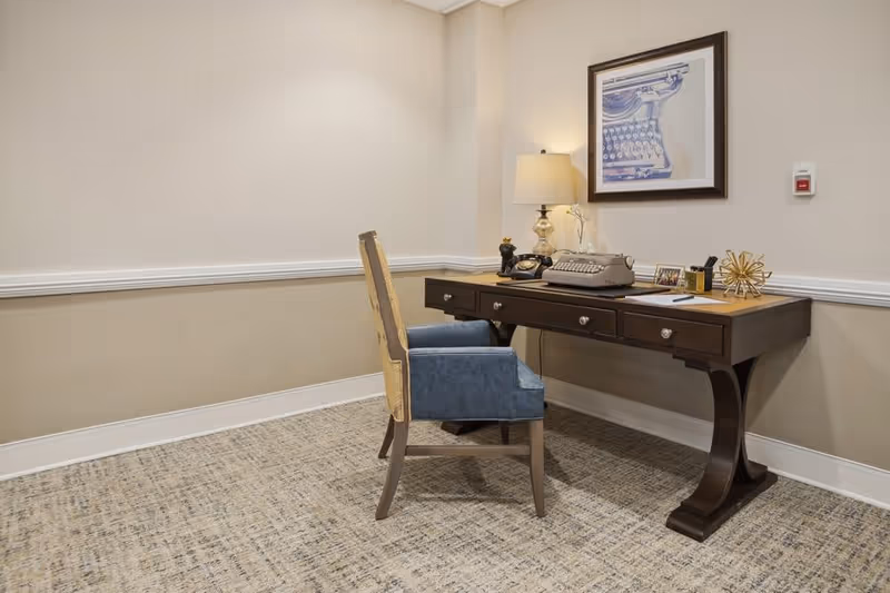 A small office corner in a room with beige walls and carpeted floor. There is a dark wooden desk with three drawers, a blue upholstered chair, a table lamp, a vintage typewriter, a black rotary phone, and decorative items on the desk. A framed picture of a typewriter hangs on the wall above the desk.