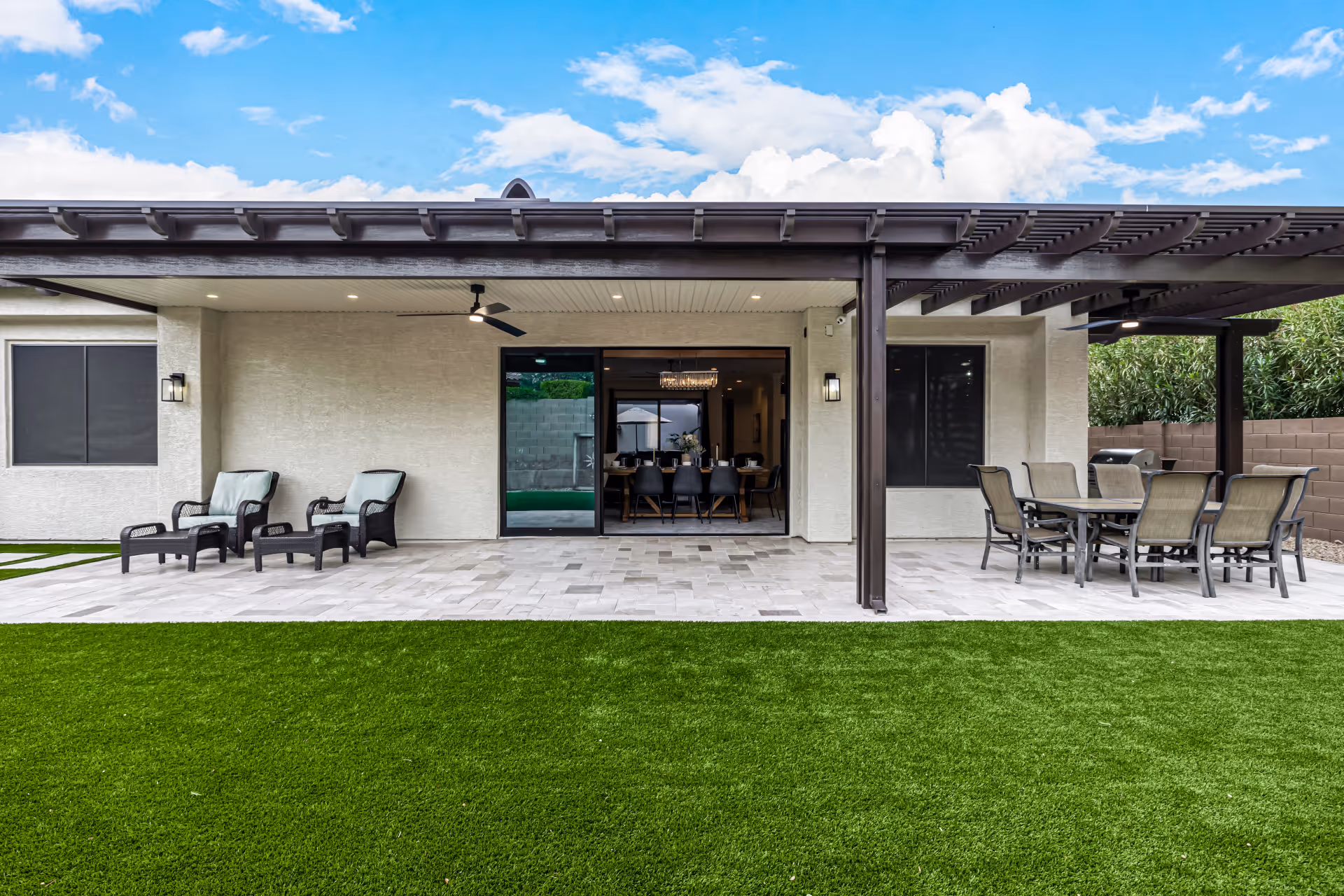 Outdoor patio area with artificial grass lawn in the foreground, two lounge chairs with footrests on the left side under a covered patio, and a dining table with six chairs on the right side under a pergola. The patio is attached to a building with sliding glass doors leading inside, showing a dining area with a chandelier.
