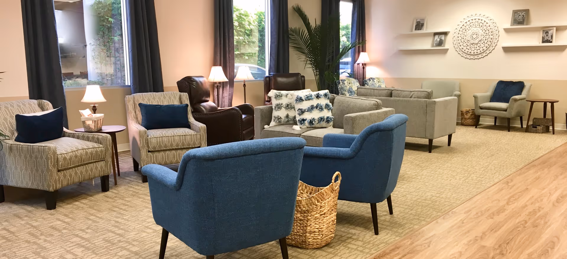 A cozy and well-lit living room area with multiple armchairs and sofas arranged for conversation. The room features beige walls, carpeted flooring, and large windows with dark curtains. There are decorative pillows on the sofas, a woven basket on the floor, table lamps, and framed photos on shelves mounted on the wall.