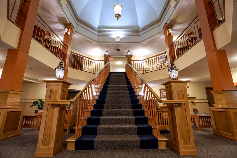 Interior view of a senior living facility featuring a wide staircase with carpeted steps and wooden railings leading to an upper floor balcony. The area is well-lit with hanging and wall-mounted lantern-style lights, wooden pillars, and beige walls. There are seating areas with wooden furniture and plants on either side of the staircase.