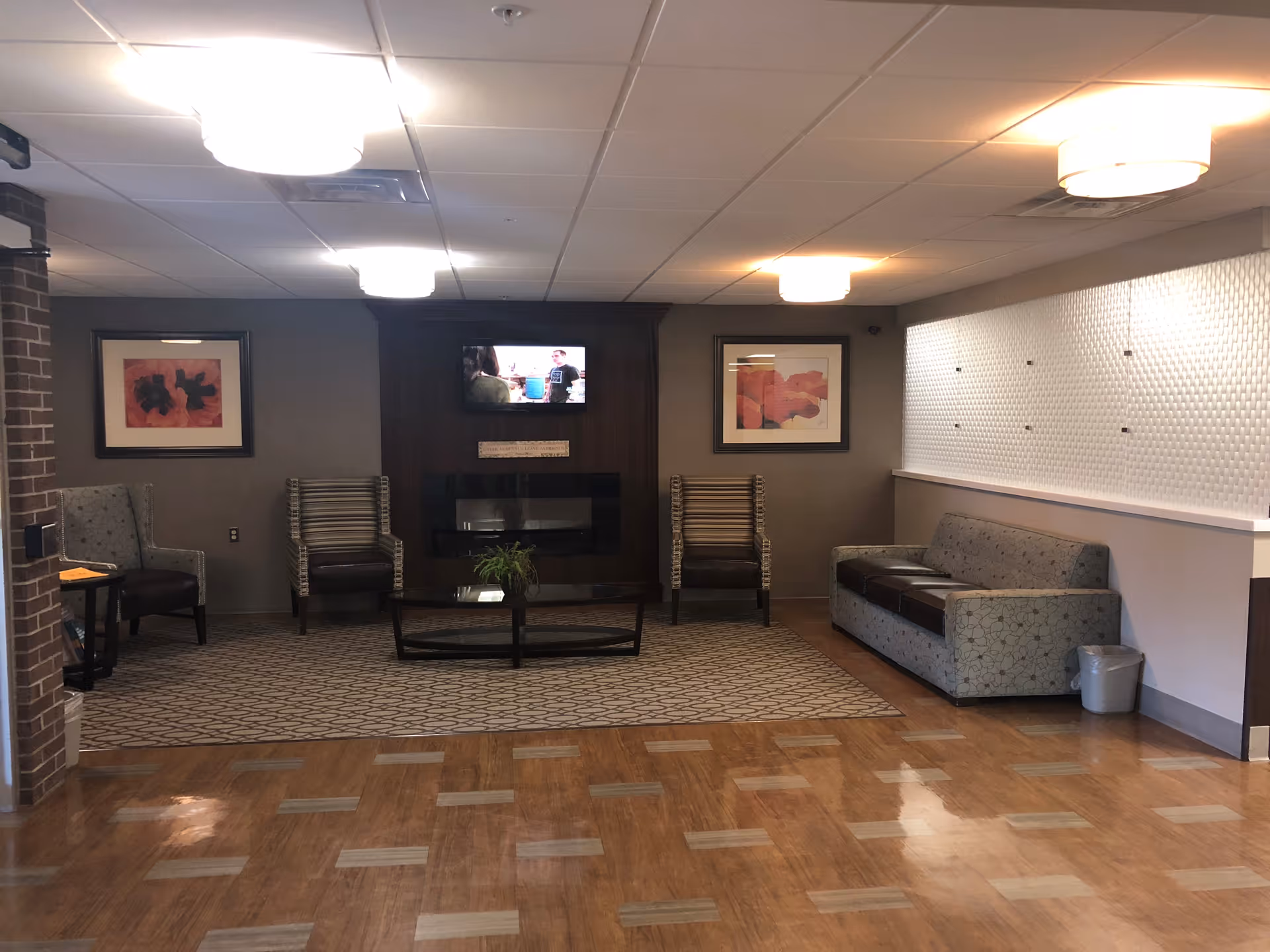 A cozy living room area with a patterned carpet, a glass coffee table, two striped armchairs, a gray sofa, and two framed floral artworks on the walls. A flat-screen TV is mounted above a modern electric fireplace on a dark wood panel wall. The room has wood flooring and ceiling lights.