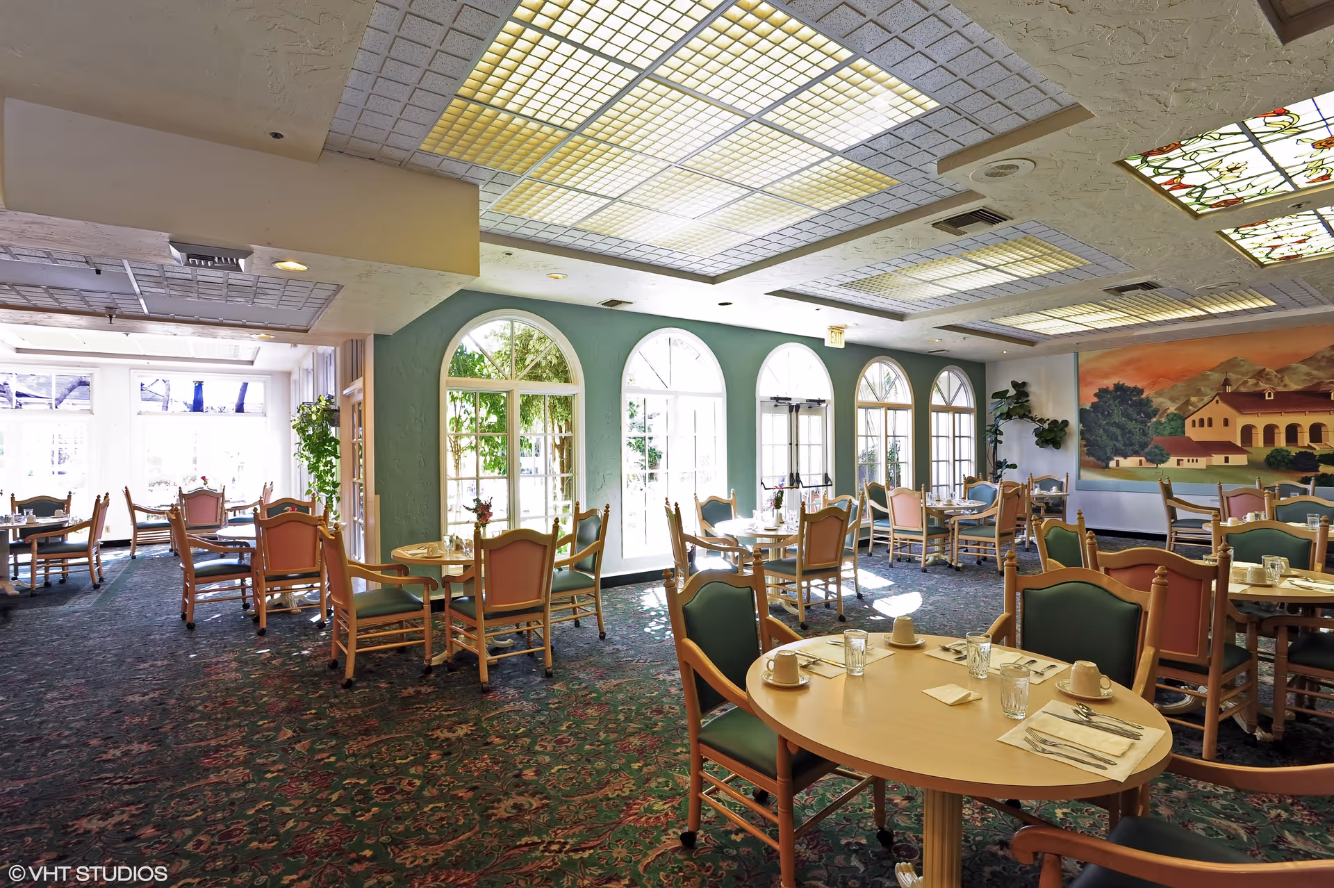 A bright dining room with multiple round tables and chairs arranged neatly. The room features large arched windows allowing natural light to fill the space, a patterned carpet, and a mural on one wall depicting a scenic landscape with a building and trees. The ceiling has decorative light panels and stained glass accents.