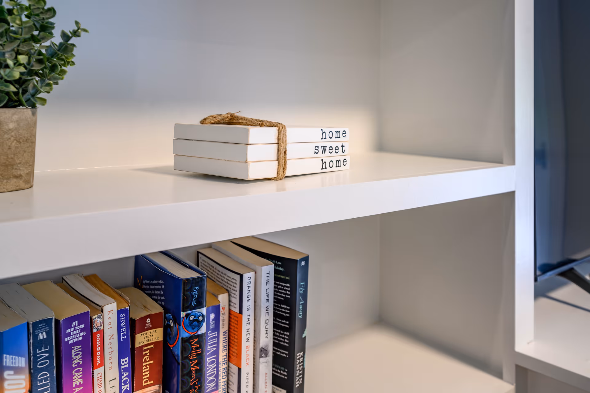 A white bookshelf with a small potted plant on the left side and a stack of three white books tied together with twine, labeled 'home sweet home'. Below the shelf, there are several books lined up vertically. The setting appears to be a cozy interior space.
