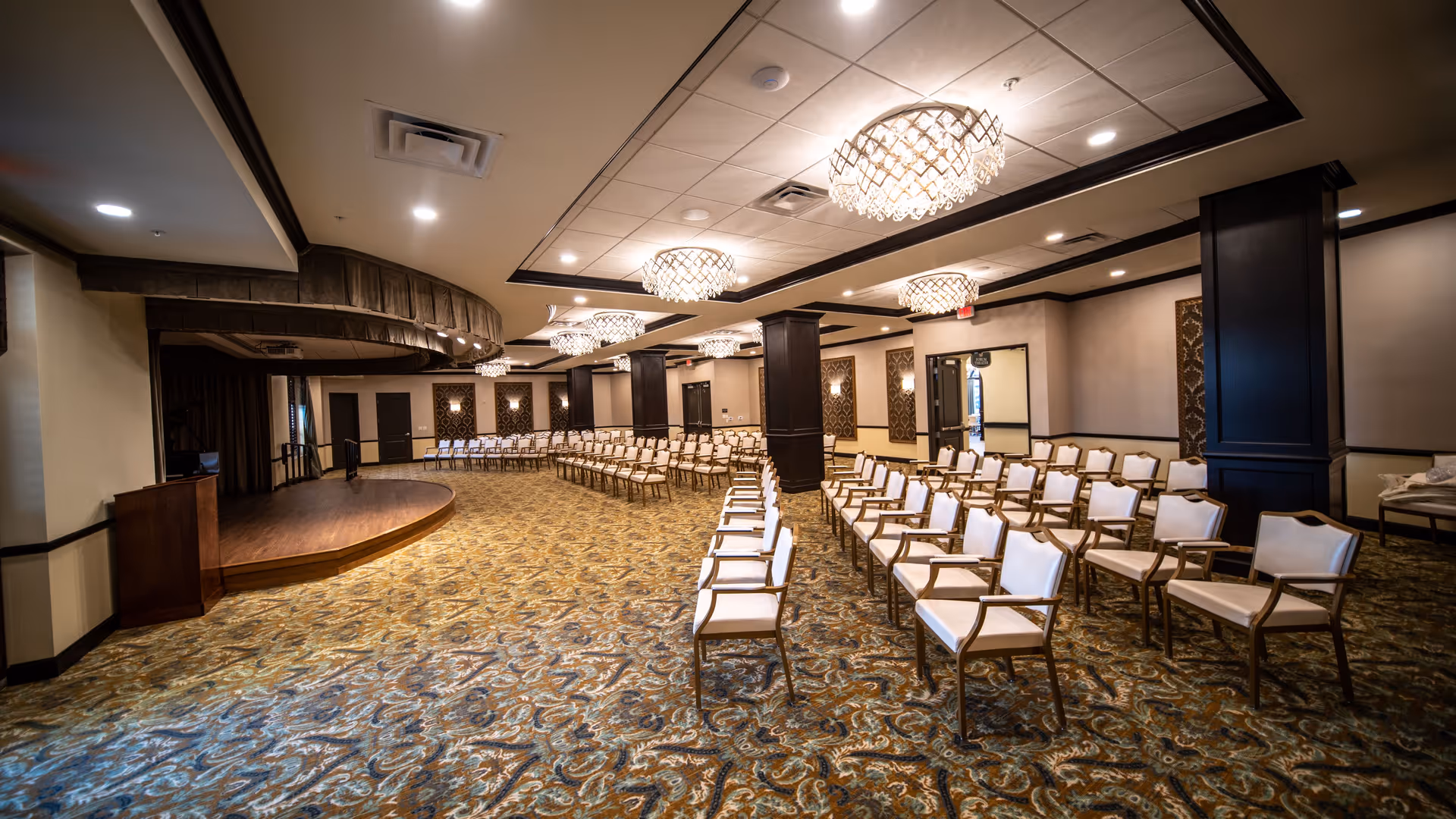 A large, elegant event room with rows of beige cushioned chairs arranged facing a small stage with a wooden podium. The room features patterned carpet, dark wood columns, and ornate chandeliers hanging from a white ceiling.