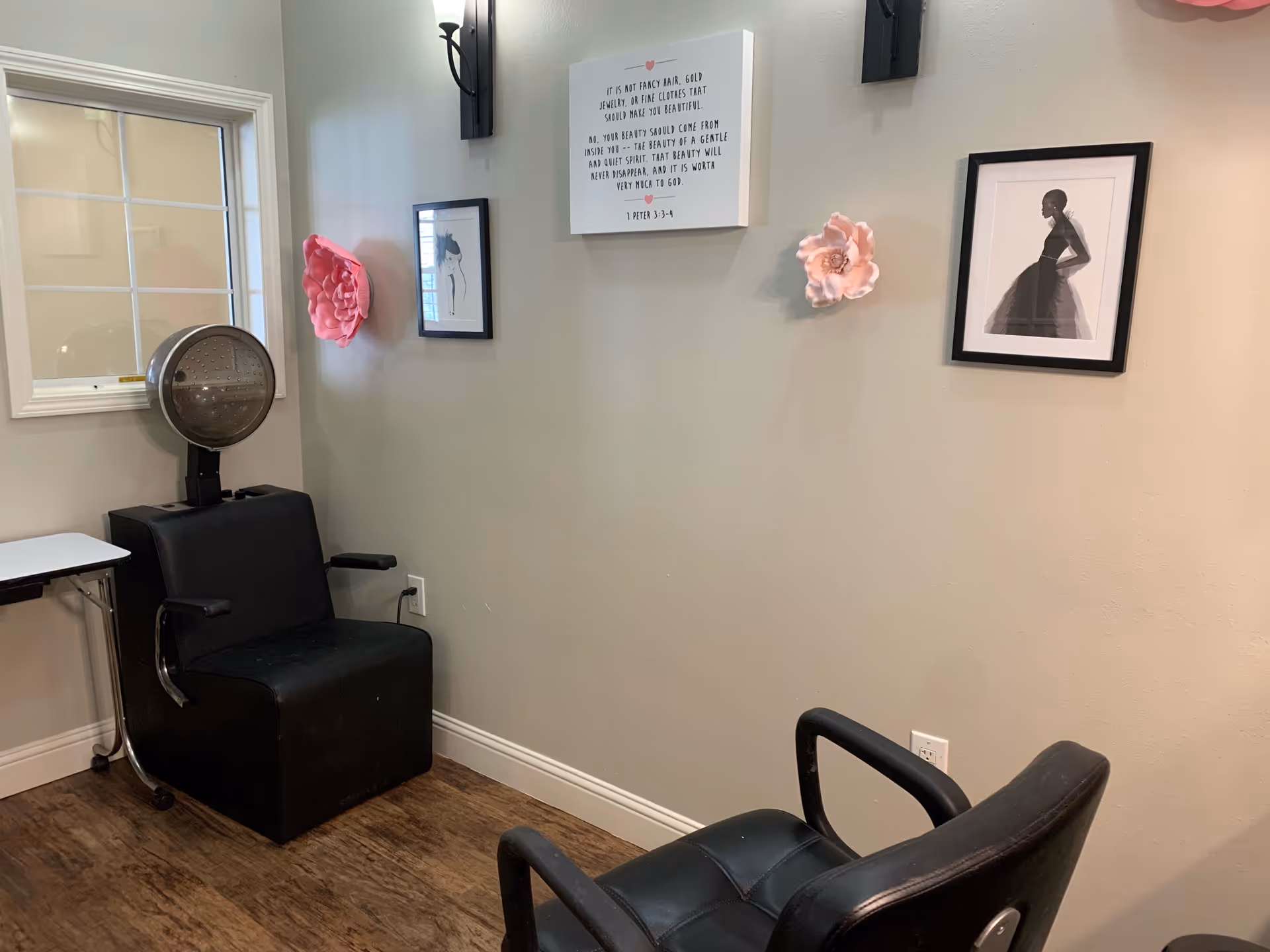 Interior view of a small salon area with two black salon chairs, one with a hair dryer hood attached. The walls are decorated with framed artwork and large pink flower decorations. There is a window on the left side and wooden flooring.
