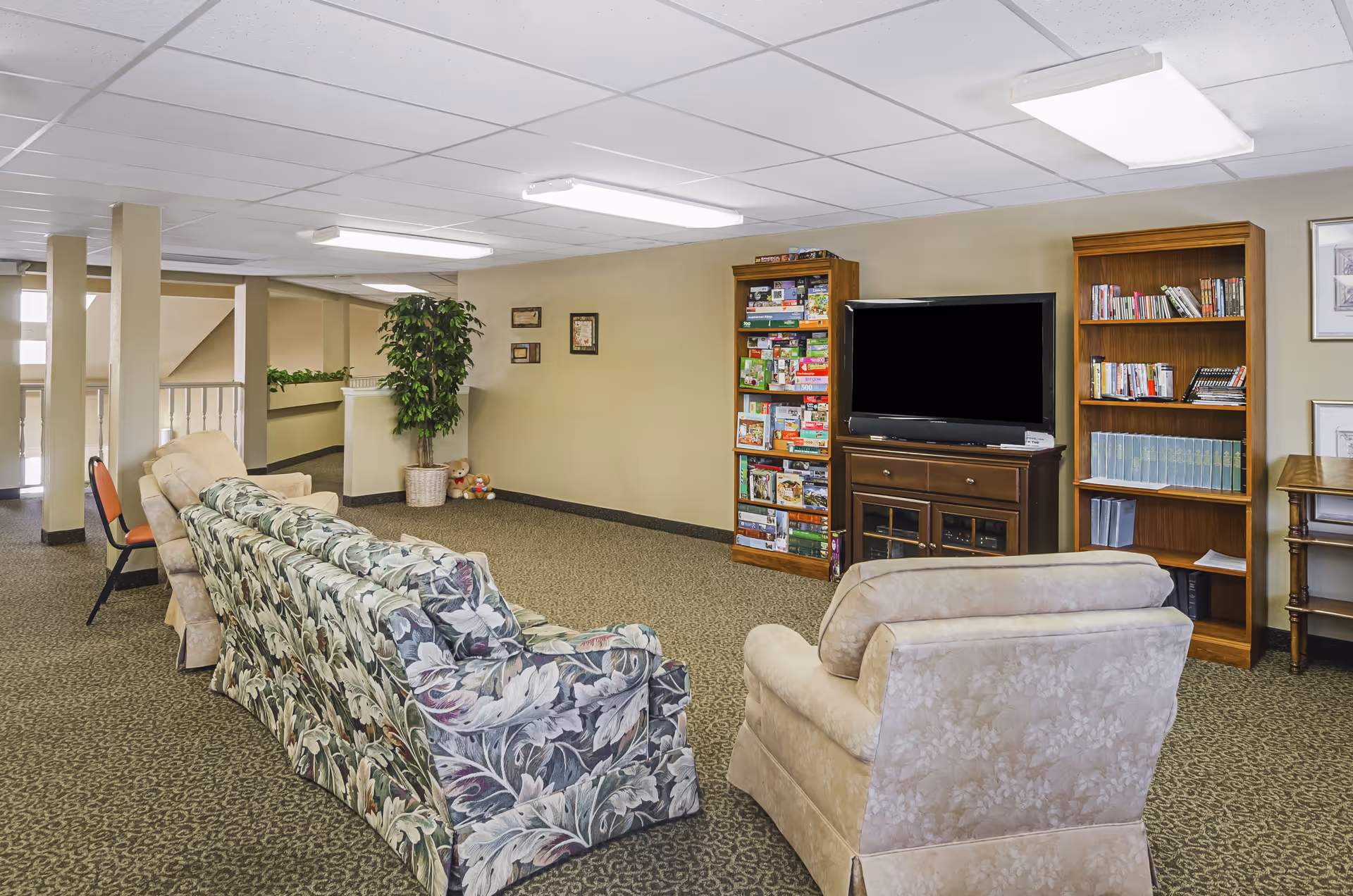 Community lounge with patterned sofas and armchairs facing a TV, bookcases and a shelf of games in a senior living facility.
