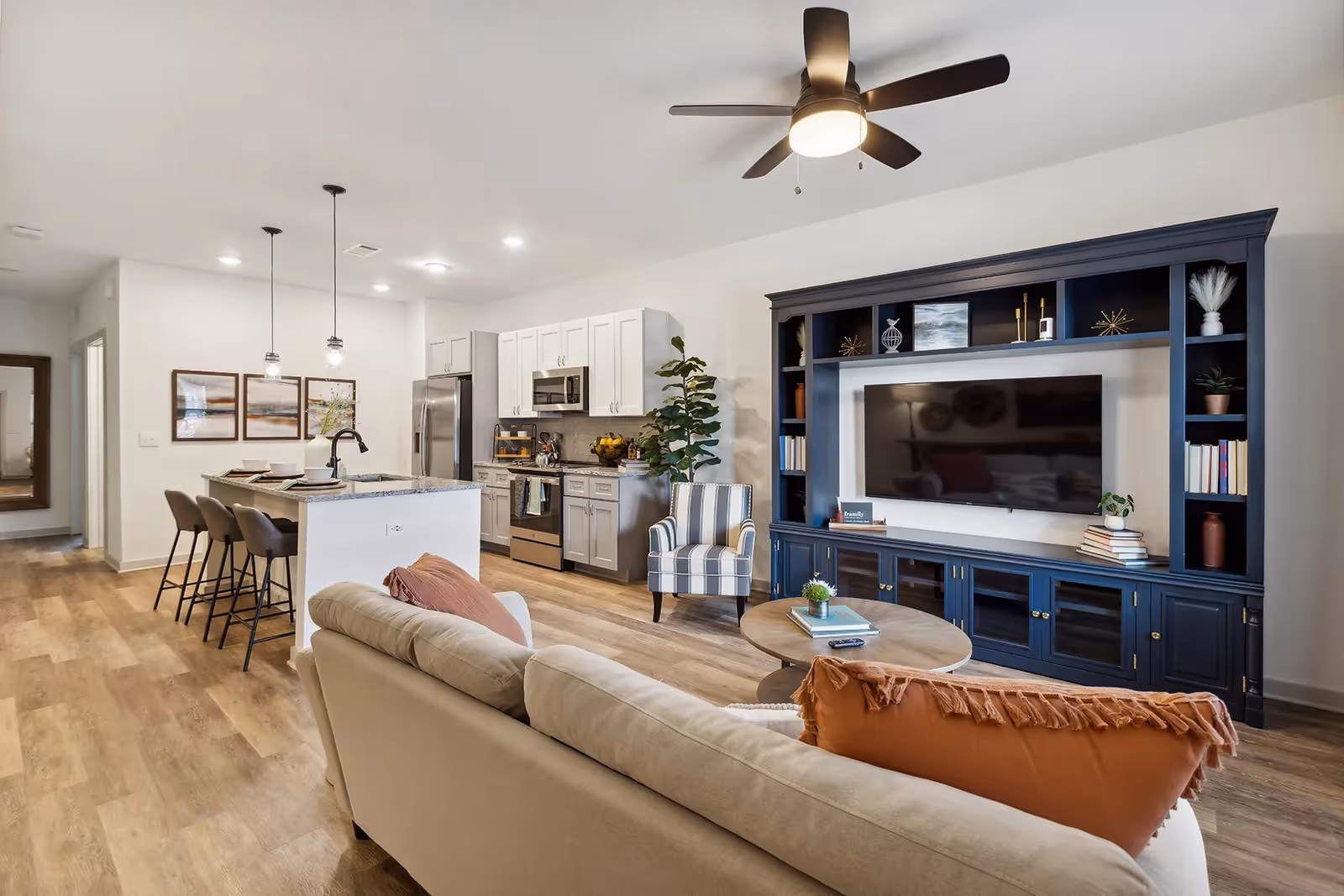 Open concept living room and kitchen area with a beige sofa adorned with orange and pink pillows, a round wooden coffee table, a striped armchair, and a large dark blue entertainment center with a flat-screen TV. The kitchen features white cabinets, stainless steel appliances, a granite countertop island with three bar stools, and pendant lighting. The room has wood flooring and a ceiling fan with lights.