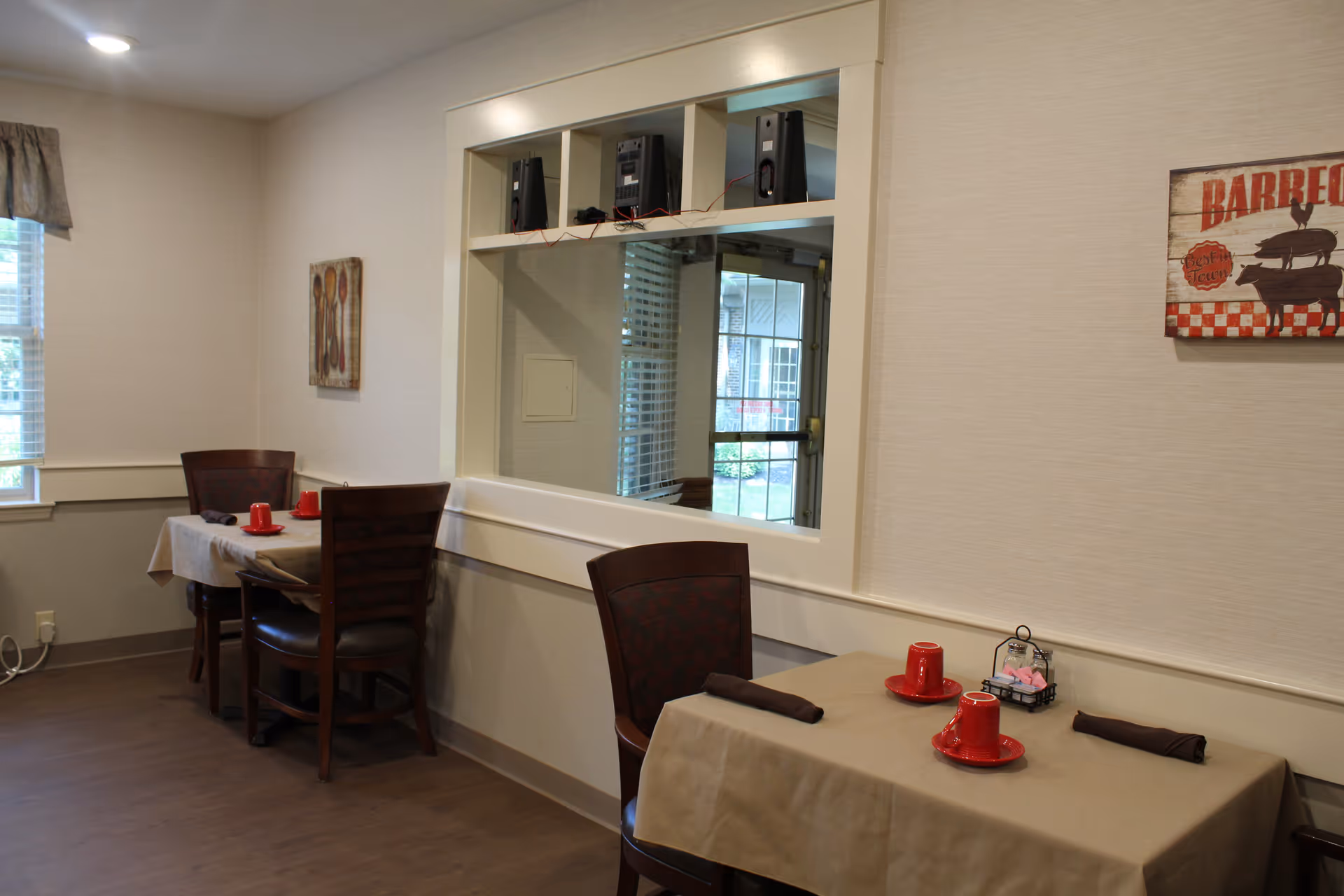 A small dining area with two tables covered in beige tablecloths, each set with two red cups and saucers, and dark brown napkins. The room has light-colored walls with two pieces of wall art, one depicting utensils and the other a barbecue theme. There is a window with blinds and a curtain on the left side, and a large interior window with shelves above it showing speakers and a view into another room.