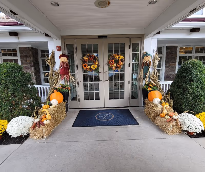 Entrance of Arbor Terrace Willistown decorated for fall with two scarecrows, pumpkins, gourds, corn stalks, and white and yellow chrysanthemums on either side of double glass doors with autumn wreaths.