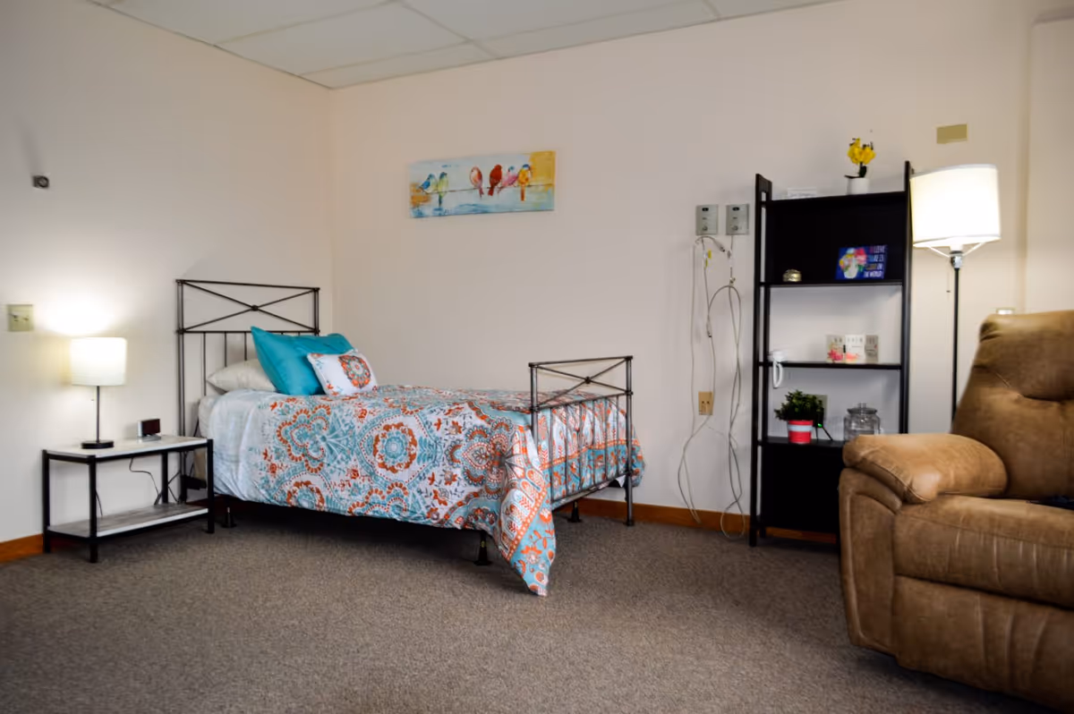 Single-occupancy room with a patterned twin bed, nightstand lamp, shelving unit, and a recliner.