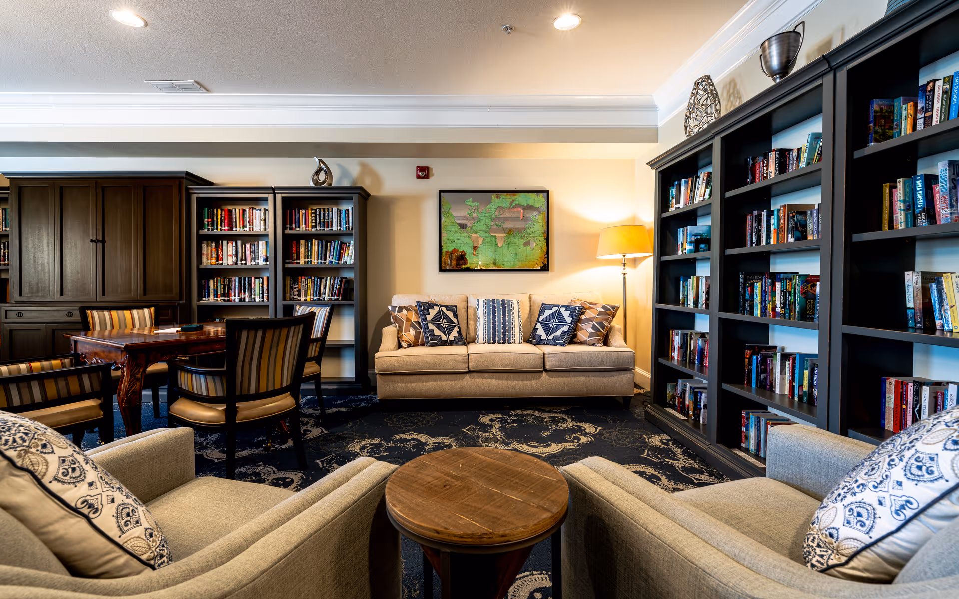 Cozy library-style lounge with bookshelves lining the walls, a sofa, armchairs, a round coffee table, and a floor lamp.