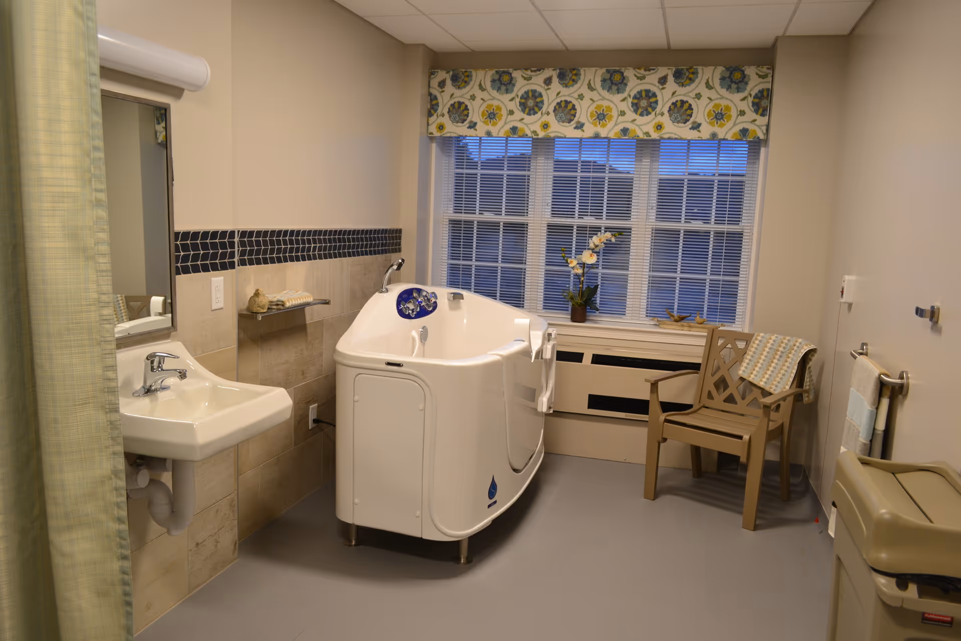 A clean and accessible bathroom featuring a walk-in bathtub with controls on the side, a white sink with a mirror above it, a chair with a folded towel on it, a window with blinds and a floral valance, and a small shelf with decorative items. The room has beige walls and tiled lower walls with a dark blue tile border.