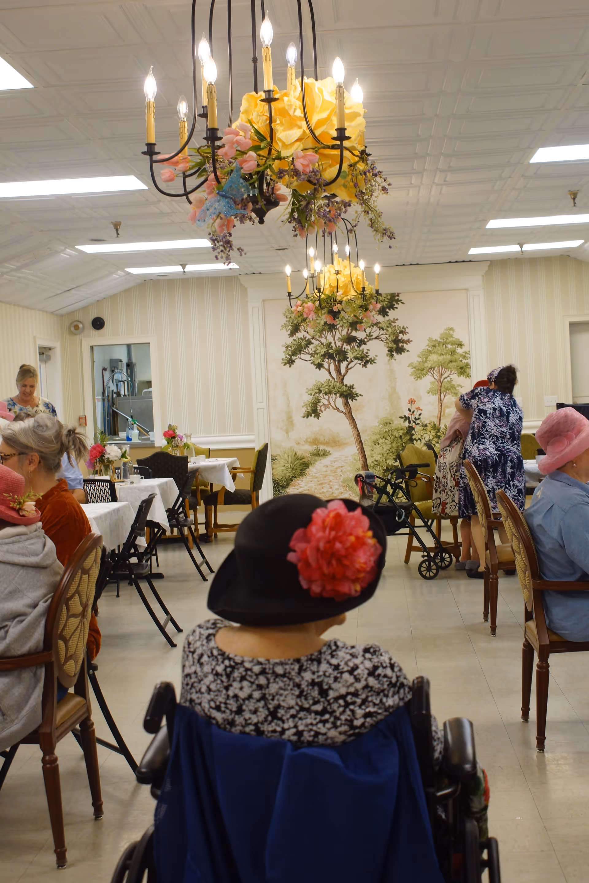 Interior of a senior living facility dining room with several elderly residents seated at tables. One resident in a wheelchair with a black hat adorned with a large red flower is in the foreground. The room features floral chandeliers, wallpaper with a nature mural, and tables covered with white tablecloths and small flower arrangements.