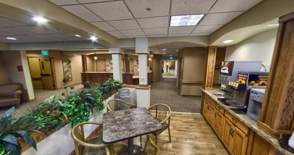 Interior view of a senior living facility common area with a small table and chairs in the foreground, a coffee and beverage station on the right, plants along a half wall, and a hallway leading to other rooms in the background.
