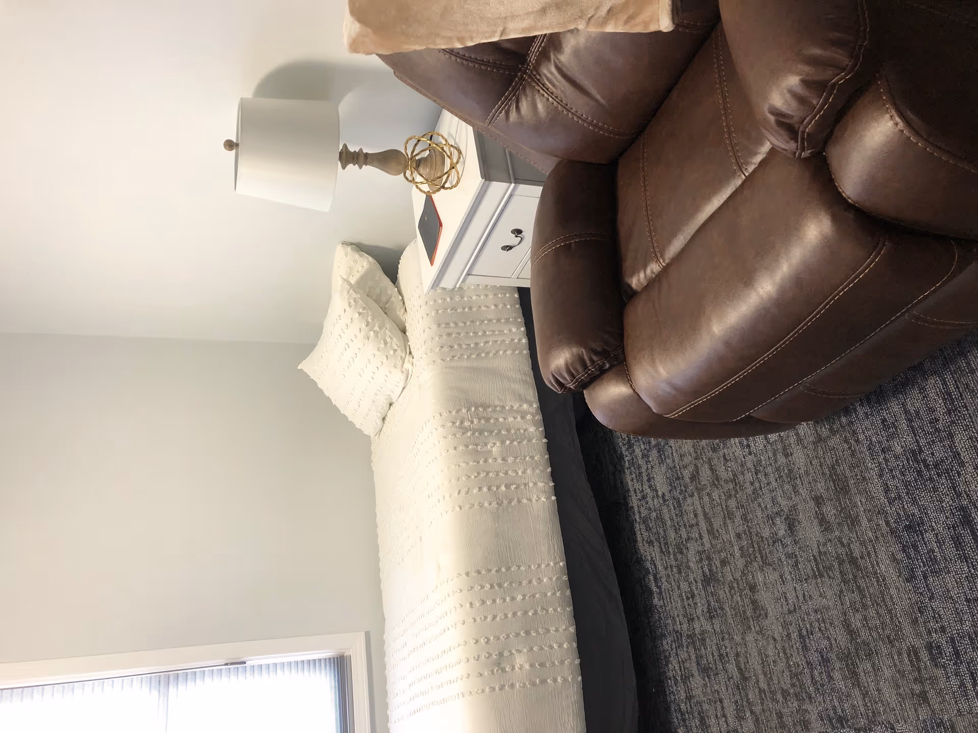 A cozy bedroom featuring a neatly made bed with white textured bedding and two pillows, a white nightstand with a decorative lamp and a small golden orb, and a brown leather recliner chair on a gray patterned rug.