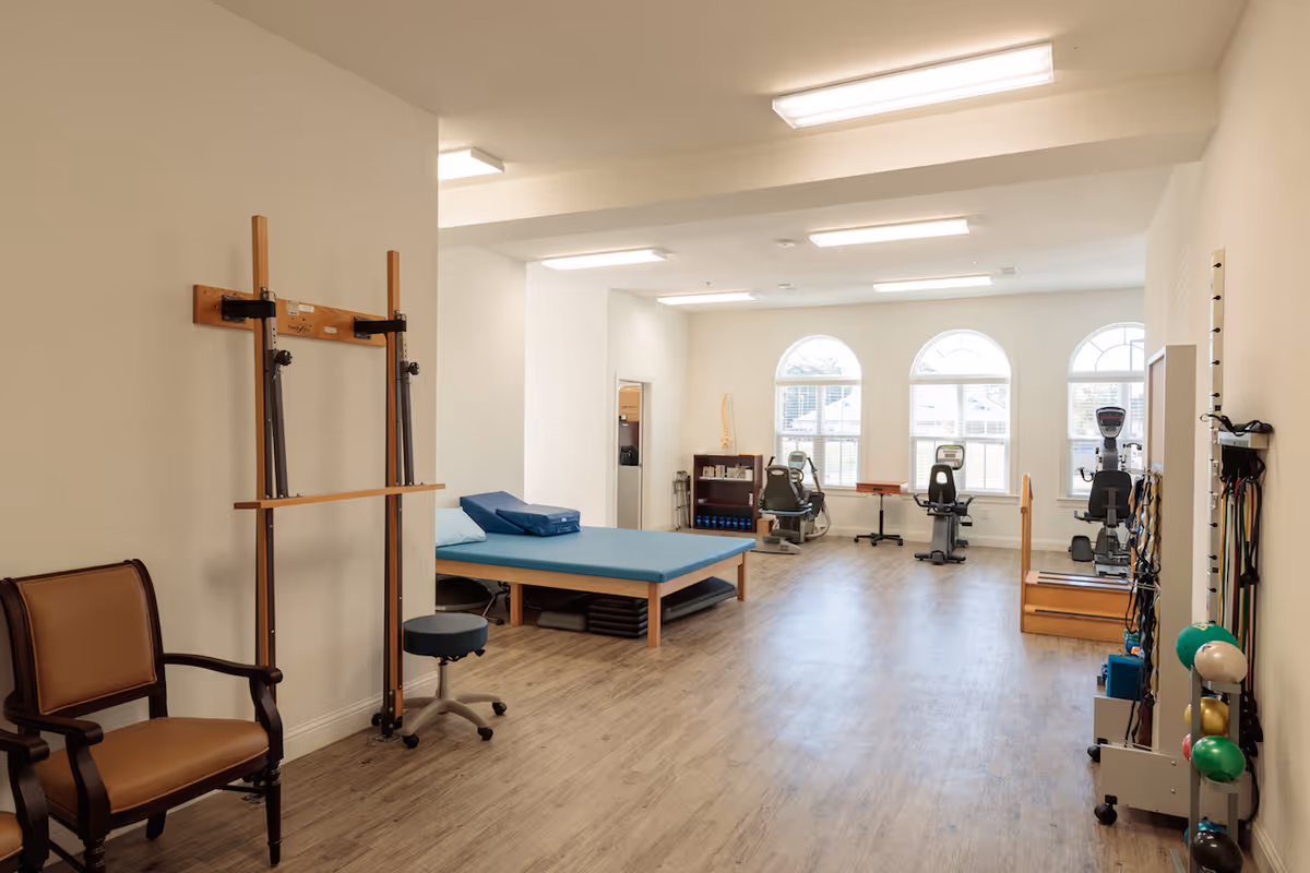 A bright physical therapy or rehabilitation room with wooden flooring, exercise equipment including stationary bikes, therapy balls, resistance bands, a padded therapy table with blue cushions, and a wooden chair. The room has three large arched windows letting in natural light.