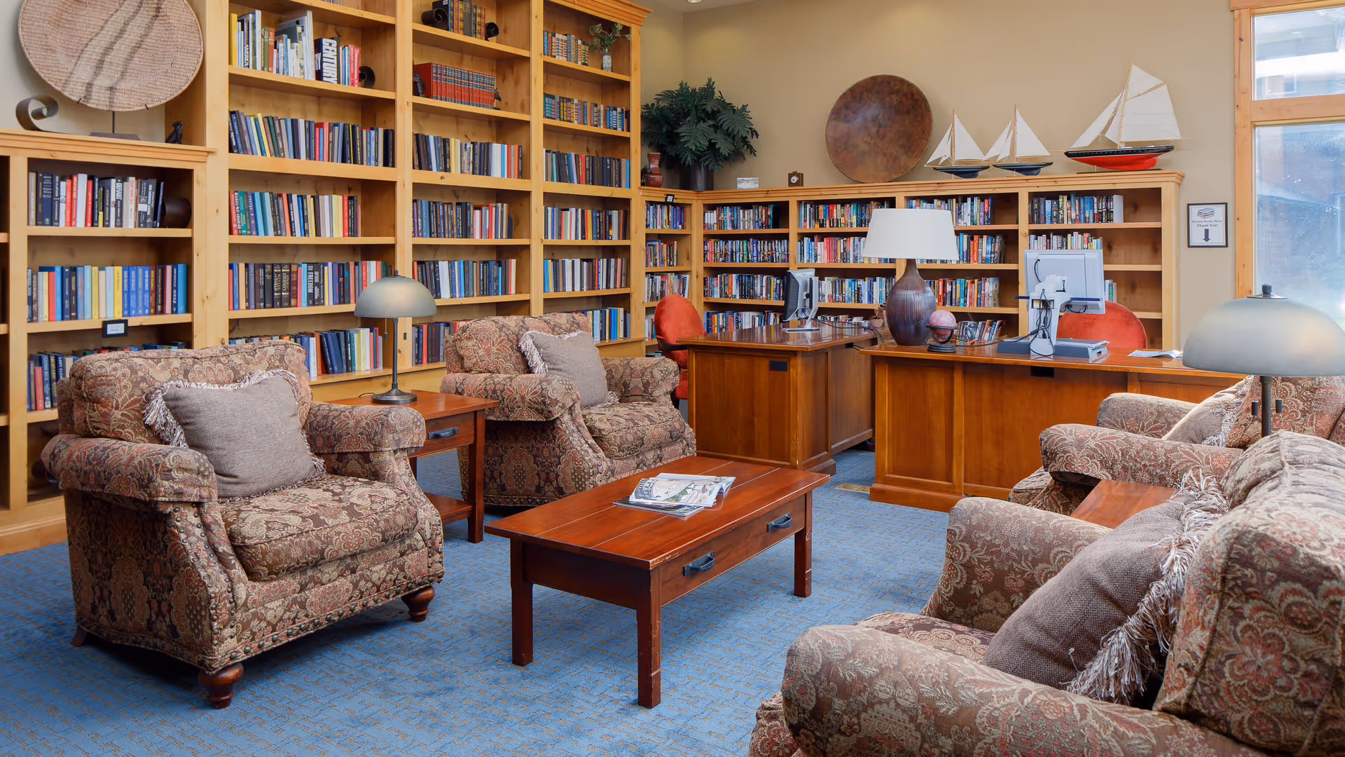 Cozy library-style lounge with patterned armchairs and sofas arranged around a wooden coffee table, built-in bookshelves, and desks with computers.