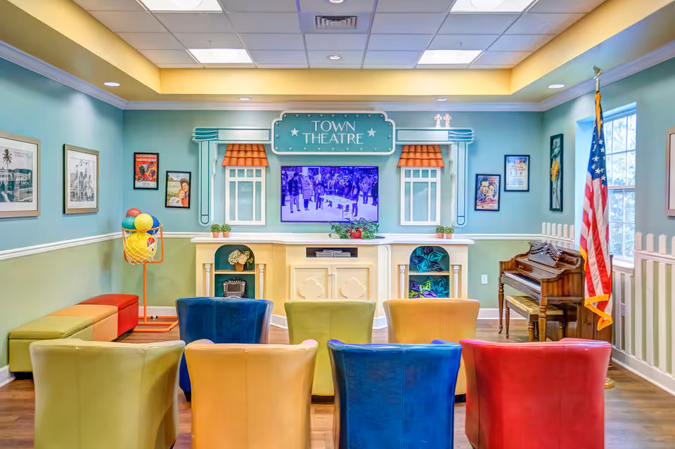 Colorful community theater room with rows of upholstered chairs facing a TV under a 'TOWN THEATRE' sign, with a piano and an American flag to the right.