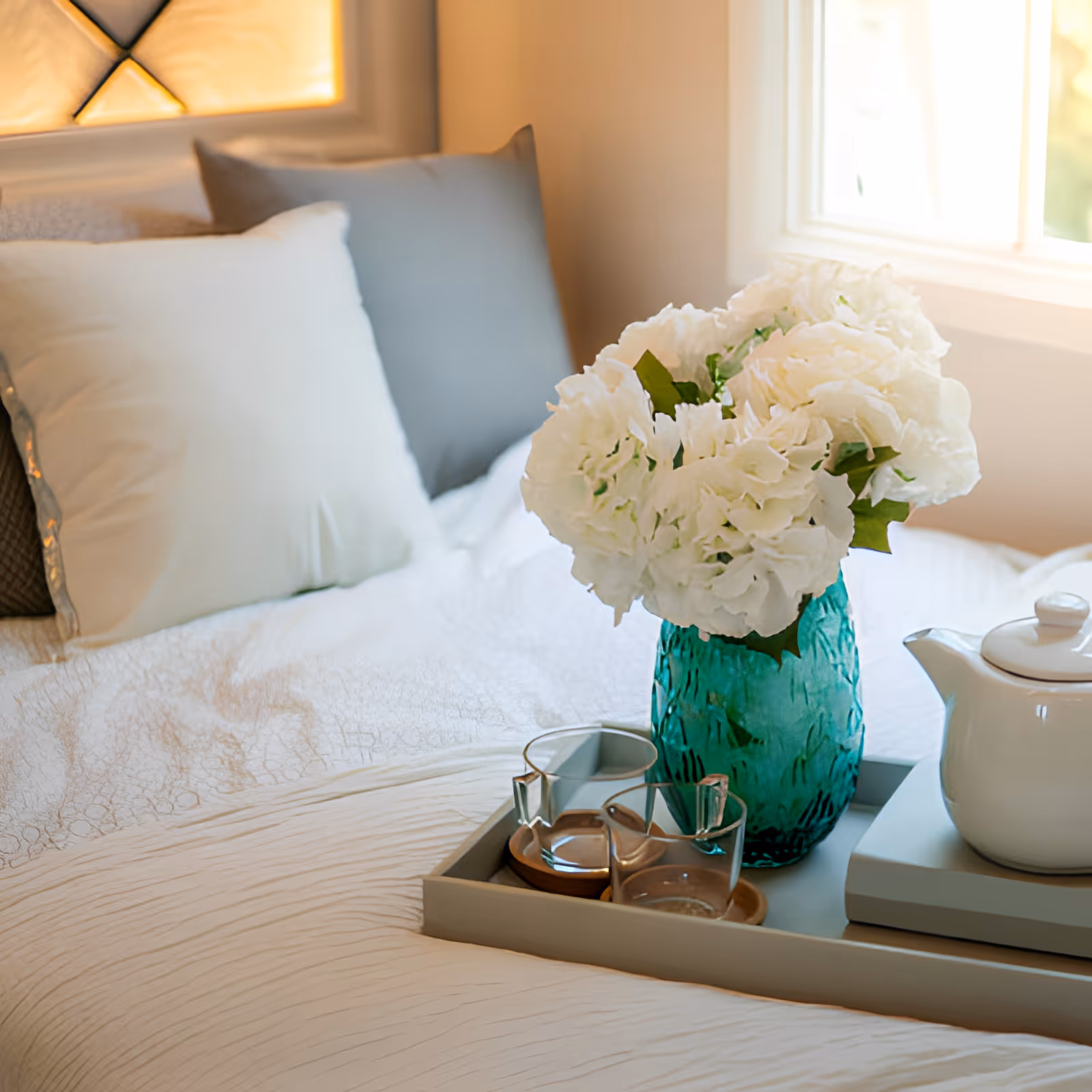 A neatly made bed with white and gray pillows, a tray on the bed holding a green vase with white flowers, two glasses, and a white teapot near a window with soft natural light.