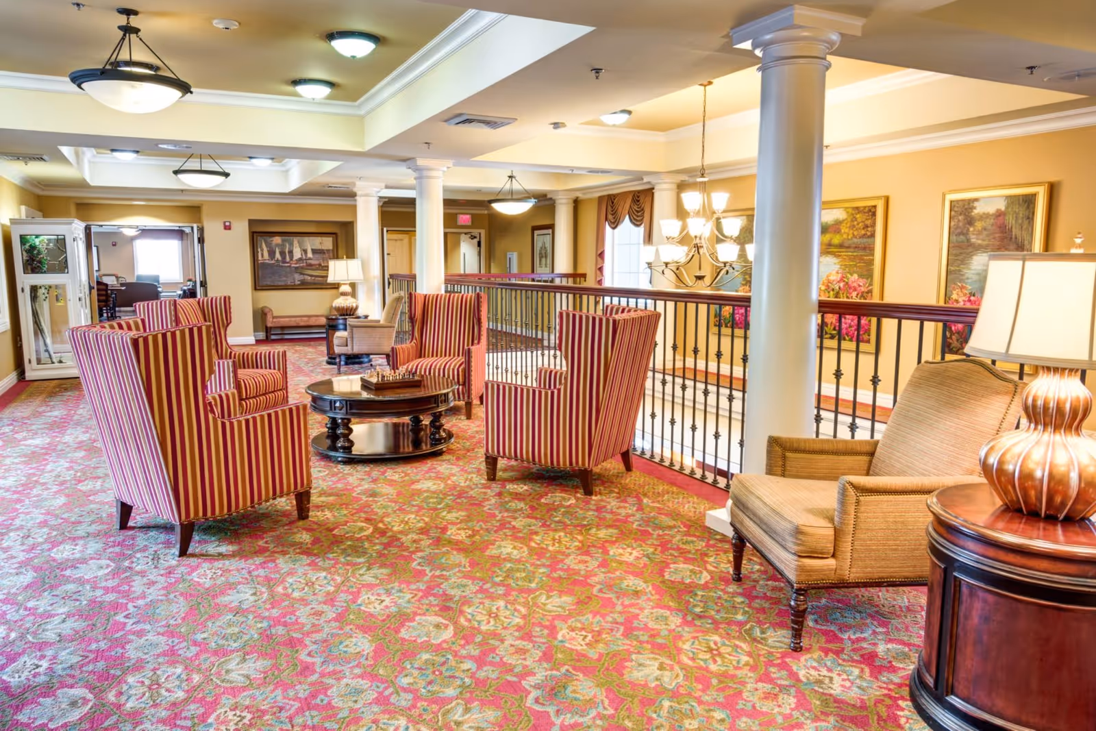 Seating area with striped armchairs, side chairs, a round coffee table, lamps and columns in a carpeted senior living lounge overlooking a railing.