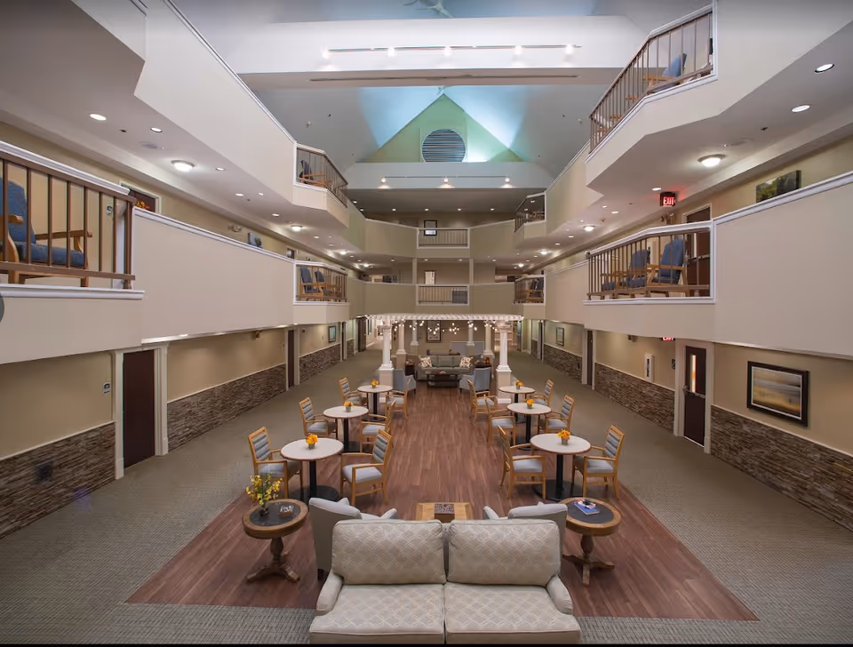 Spacious, well-lit common area in a senior living facility with multiple round tables and chairs arranged on a wood floor section, surrounded by carpet. Comfortable seating areas with sofas and armchairs are visible, along with a two-story balcony overlooking the space. The ceiling is high with a triangular window letting in natural light.