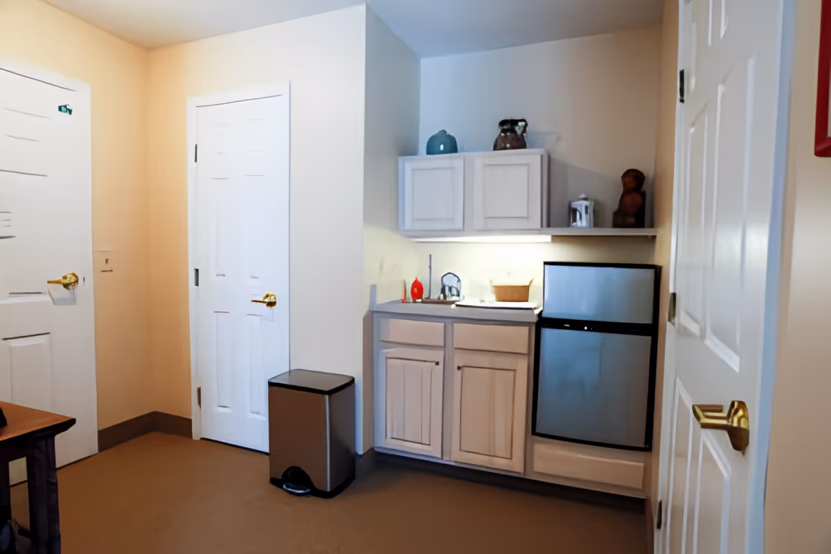 Small kitchenette area with upper and lower cabinets, a sink, a mini refrigerator, and a trash can beside two closed doors.