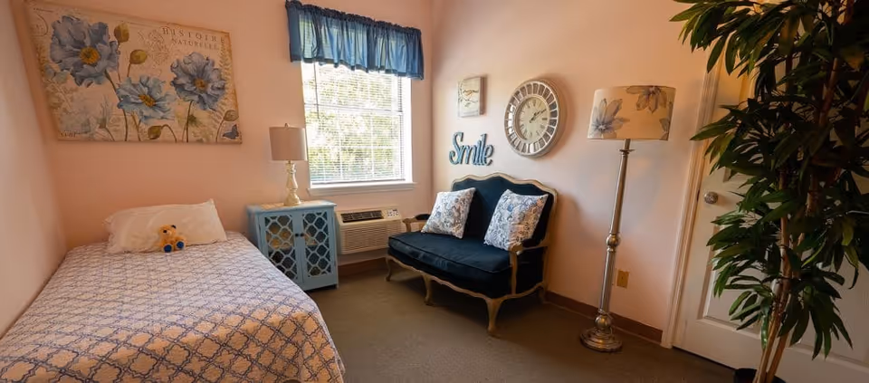 A cozy bedroom in an assisted living facility featuring a single bed with a patterned blue and white bedspread and a small stuffed animal on the pillow. Next to the bed is a blue nightstand with a lamp. A window with blue curtains lets in natural light. Across from the bed is a dark blue loveseat with two patterned pillows, a wall clock, and decorative wall art including the word 'Smile'. A floor lamp with a floral lampshade and a tall green plant are also visible.