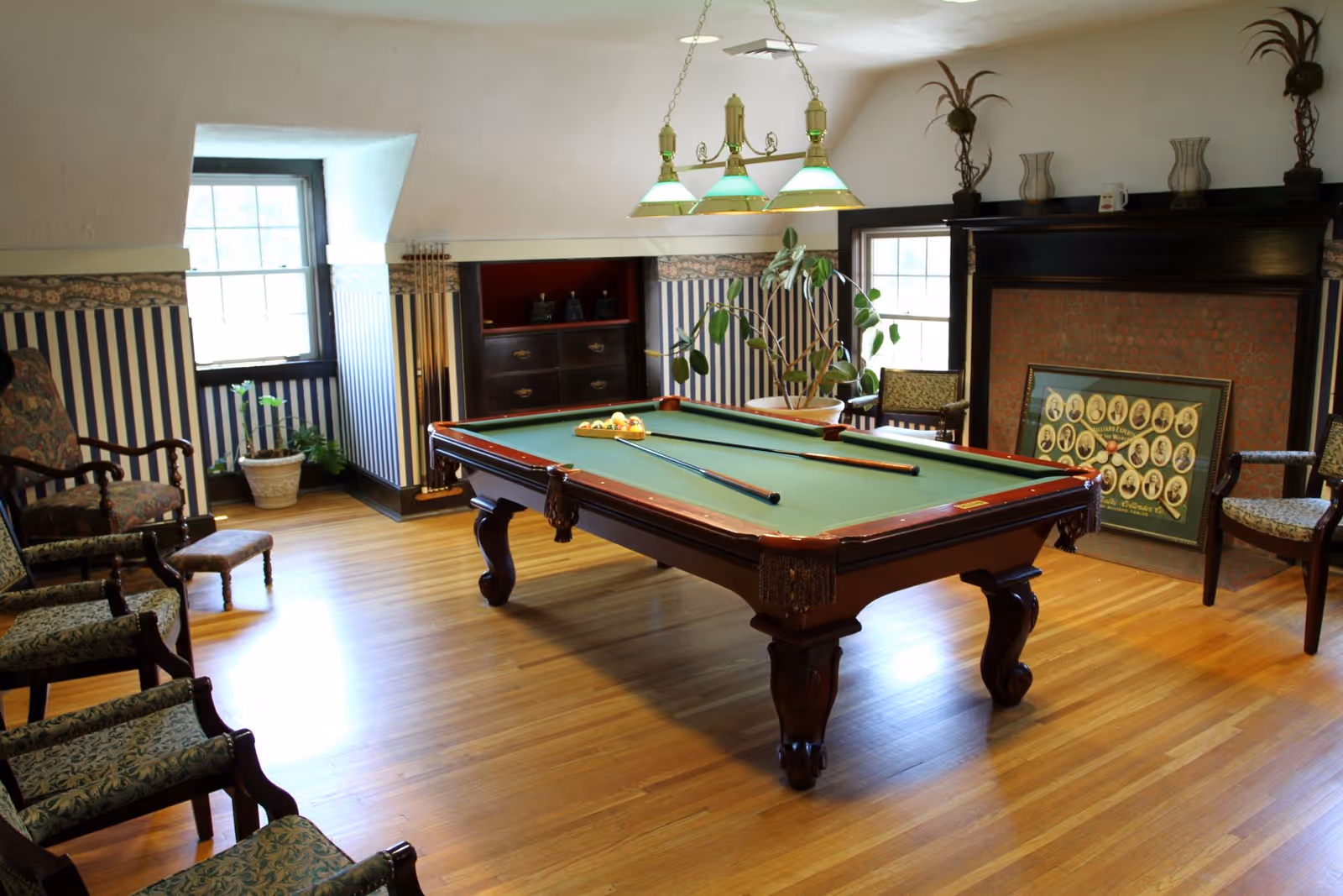 A well-lit room with a wooden pool table in the center, surrounded by several upholstered chairs. The room has hardwood floors, two windows, a fireplace with a framed photo resting on the hearth, and decorative plants on the mantel and near the windows. The walls have striped wallpaper with a floral border near the ceiling.