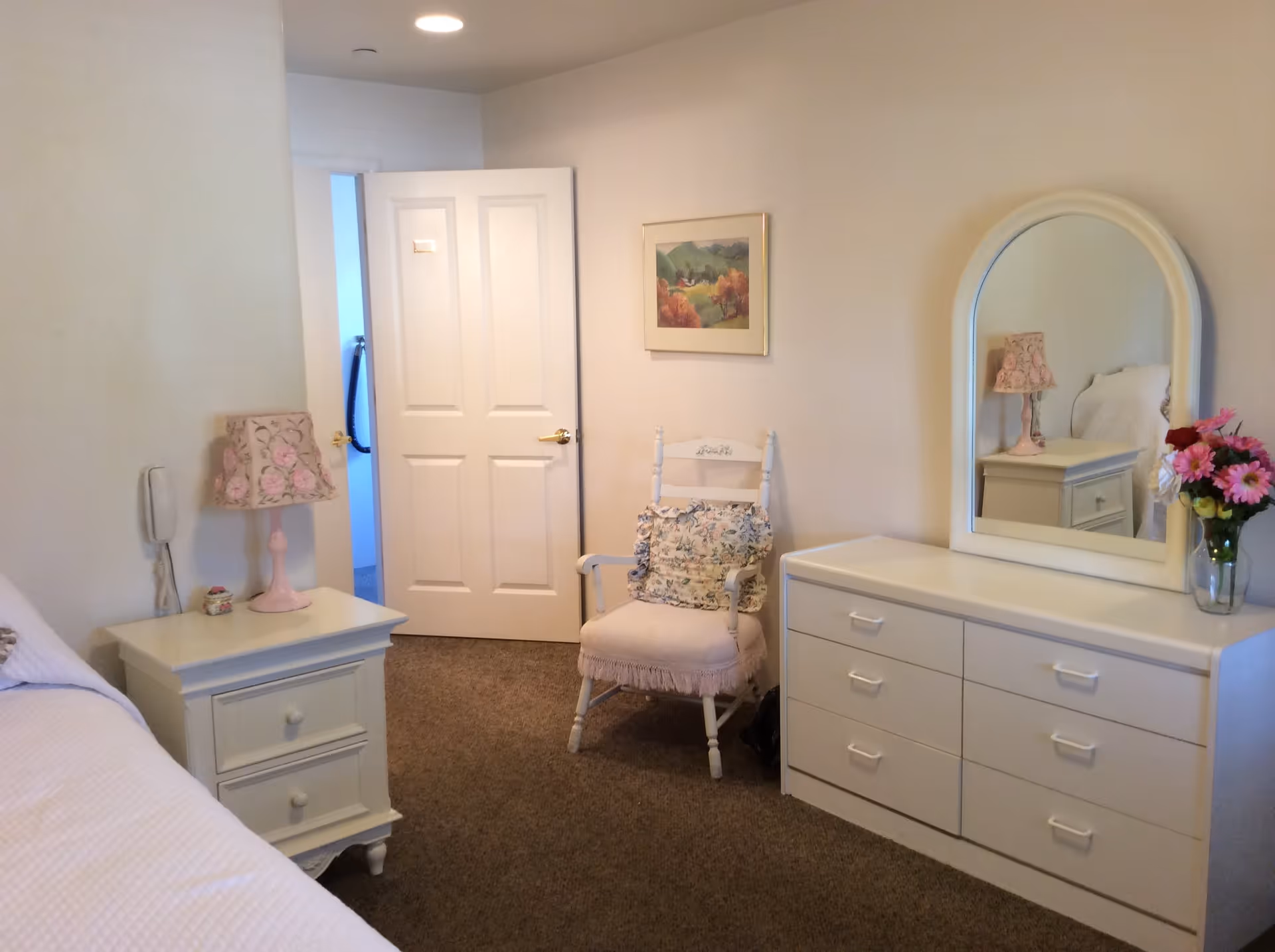 A cozy bedroom with beige walls and carpeted floor. The room features a white bed partially visible on the left, a white nightstand with a pink floral lamp, a white wooden chair with a floral cushion, and a white dresser with six drawers and an arched mirror. A vase with colorful flowers is placed on the dresser, and a framed landscape painting hangs on the wall above the chair. The door to the room is open, showing a glimpse of another room with blue walls.