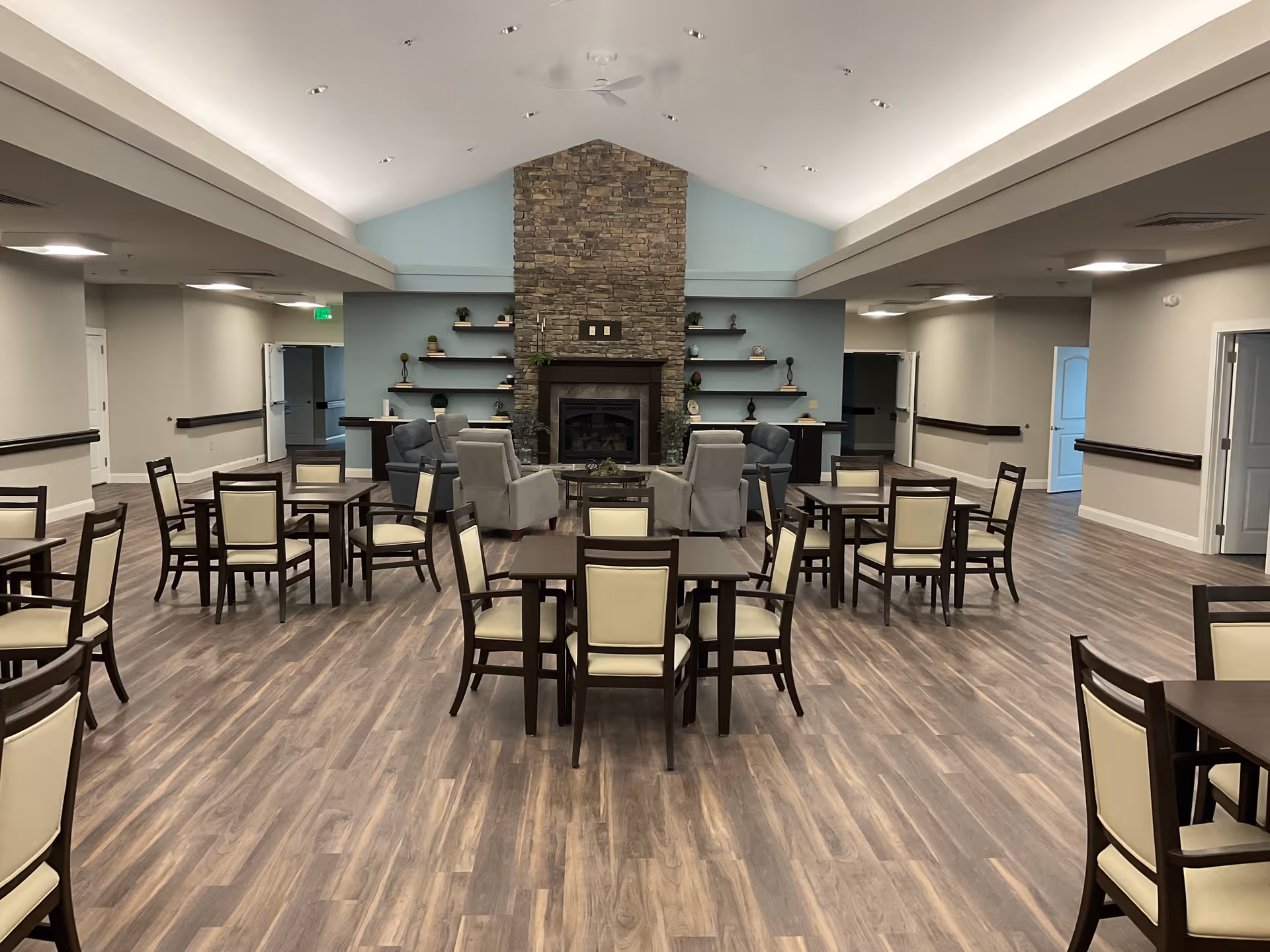 A spacious common area with multiple tables and chairs arranged on a wood floor. At the far end, there is a stone fireplace with shelves on either side decorated with small plants and ornaments. Comfortable armchairs are placed around the fireplace. The ceiling is vaulted with recessed lighting and a ceiling fan.