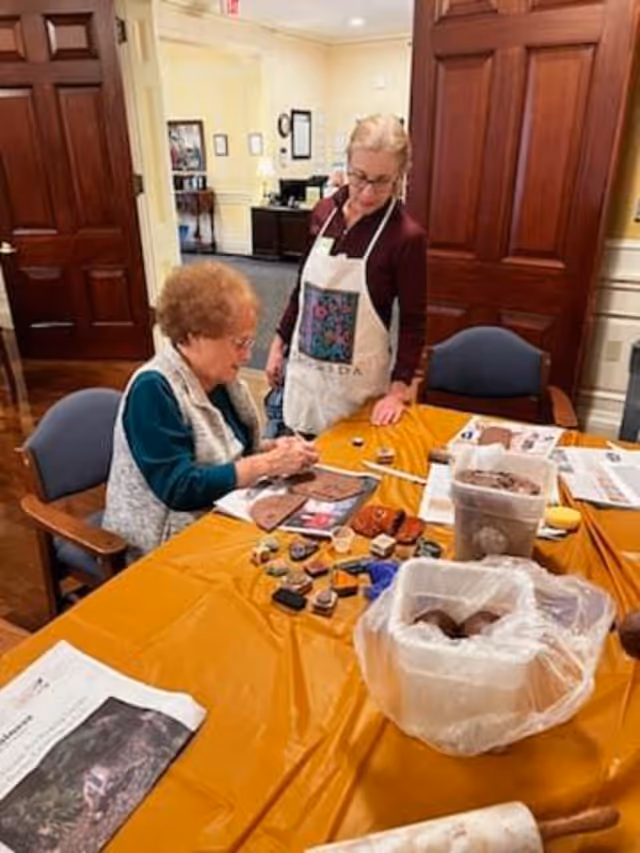 Two elderly women engaged in an arts and crafts activity at a table covered with an orange tablecloth. One woman is seated and painting on a piece of material, while the other stands beside her wearing an apron. Various art supplies and containers are spread out on the table. The room has wooden doors and a warm, inviting interior.