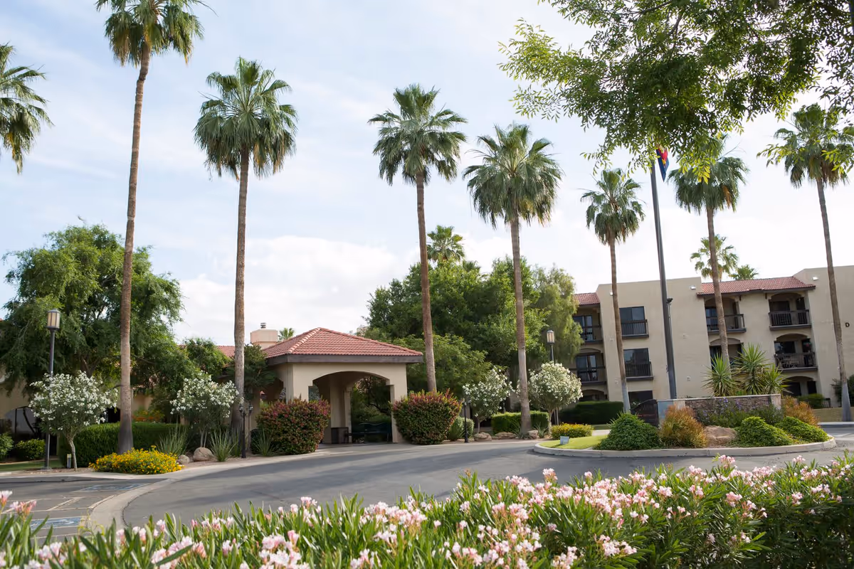 Exterior view of Sierra Winds facility showing a driveway with a covered entrance, surrounded by palm trees, flowering bushes, and other greenery under a partly cloudy sky.