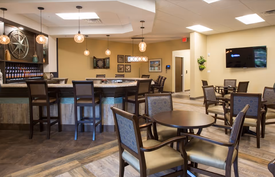 Open senior living common area with round tables and chairs, a bar with stools, pendant lights, a wall-mounted TV and a dartboard.