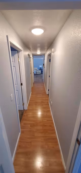 Long narrow indoor hallway with wood laminate flooring, white walls, several doorways and ceiling lights leading to a sitting area at the far end.