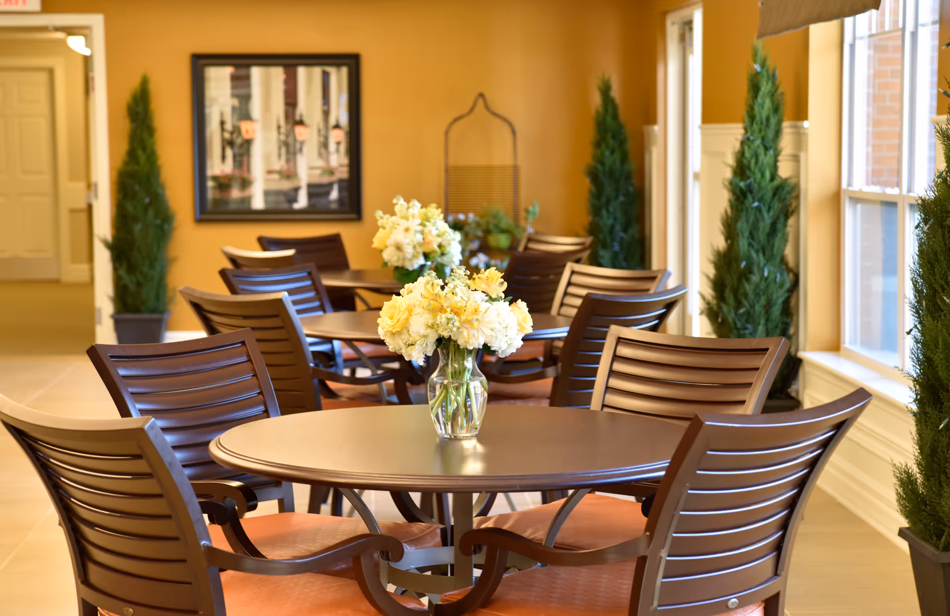 Dining area with round tables, wooden chairs, vases of yellow flowers, and potted greenery in a bright common room.