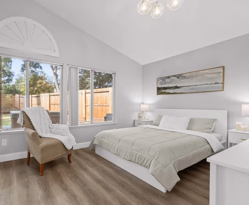 A bright and airy bedroom with a large bed covered in light-colored bedding, two bedside tables with lamps, a comfortable armchair draped with a white textured blanket, large windows showing a fenced backyard, and a landscape painting on the wall above the bed.