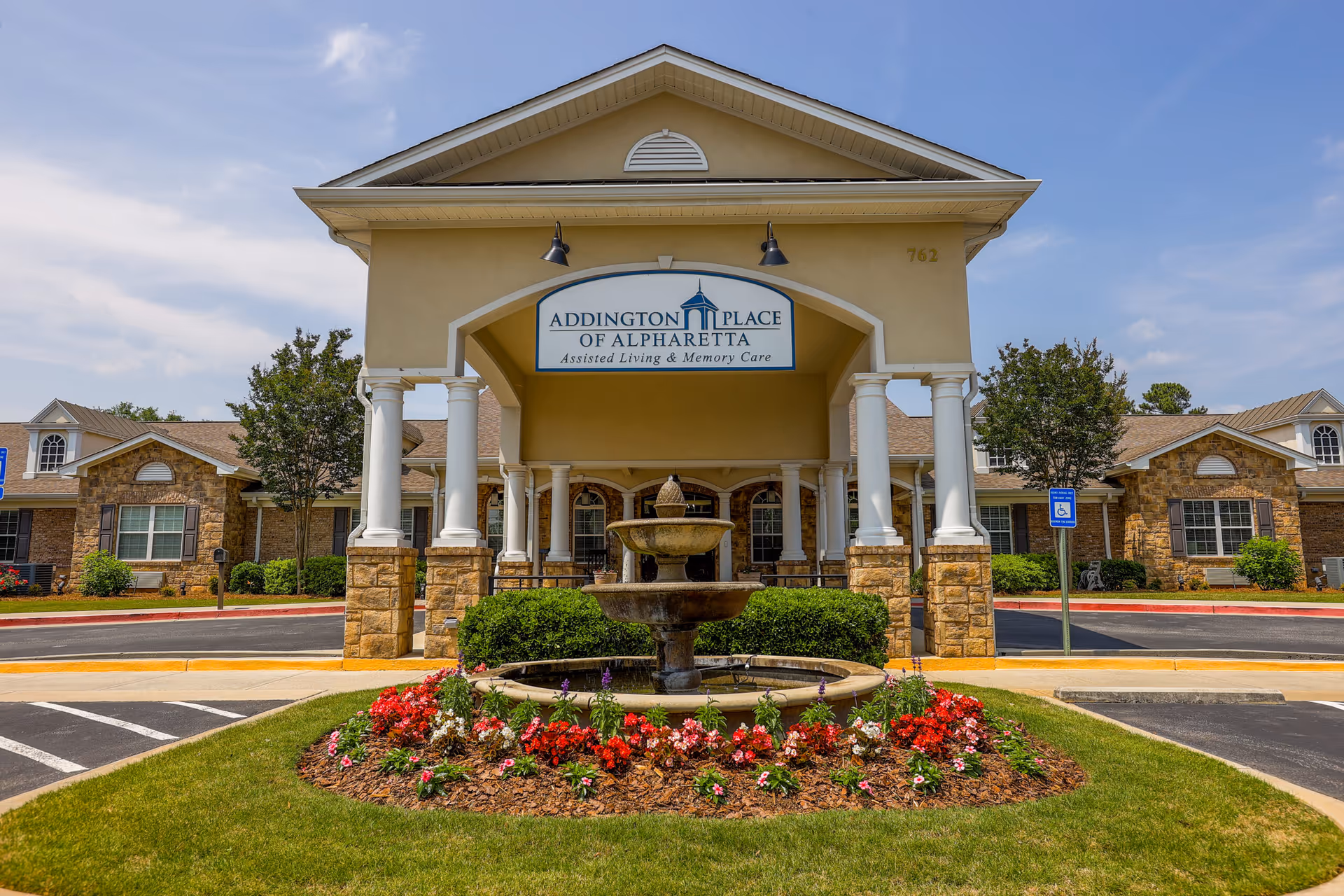 Front exterior view of Addington Place of Alpharetta assisted living and memory care facility with a covered entrance supported by white columns, a central water fountain surrounded by flowers, and a clear blue sky.