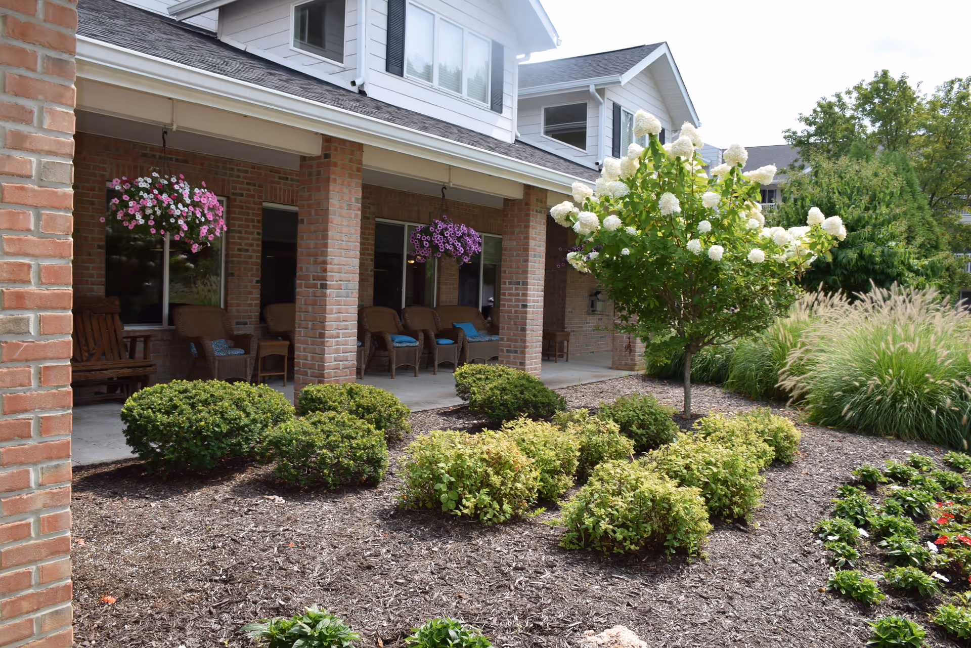 Outdoor view of a senior living facility porch with brick pillars, several chairs with cushions, hanging flower baskets, and landscaped garden beds with bushes, a small tree with white flowers, and ornamental grasses.