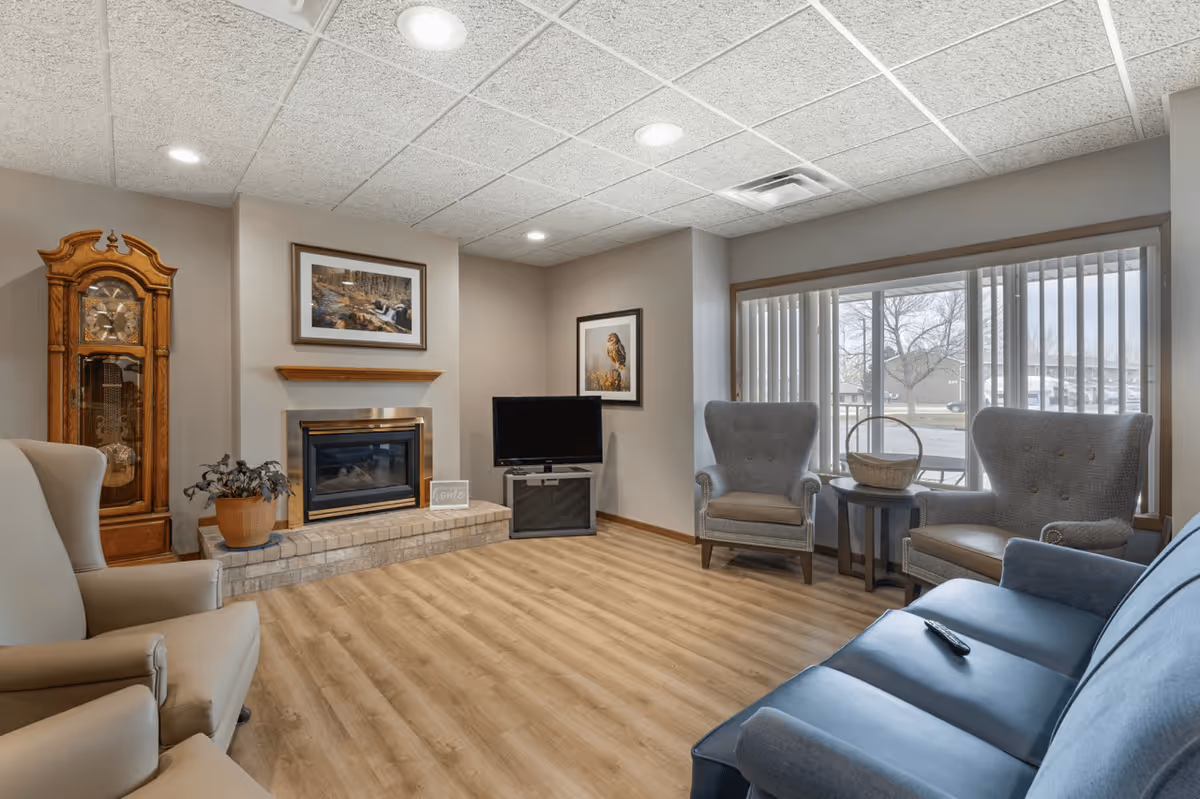 Cozy furnished common living room with a fireplace, TV, armchairs, couch, and large window overlooking the grounds.