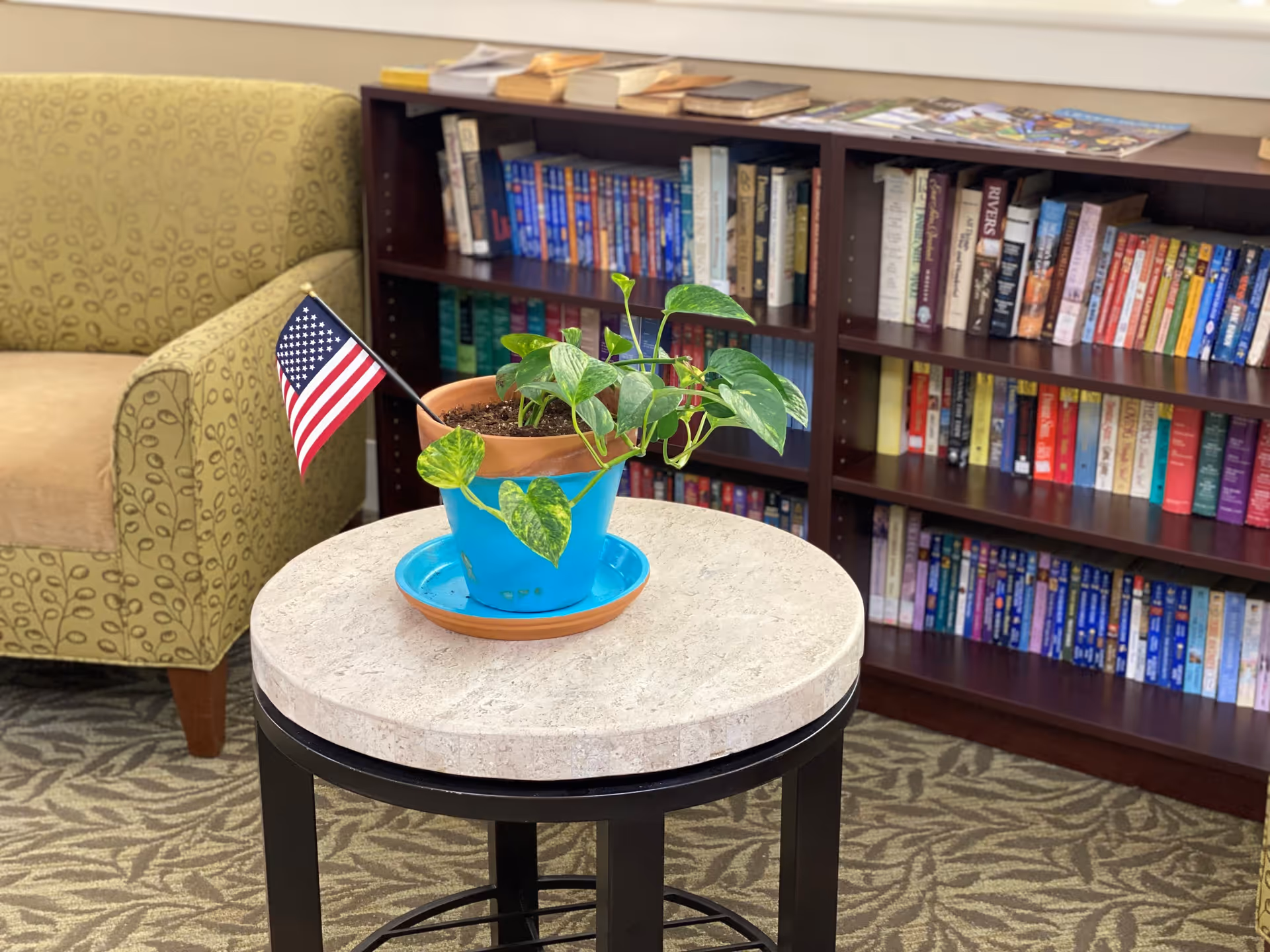A small round table with a potted plant in a blue pot and a small American flag on it. Behind the table is a green patterned armchair and a wooden bookshelf filled with books. The floor has a patterned carpet.