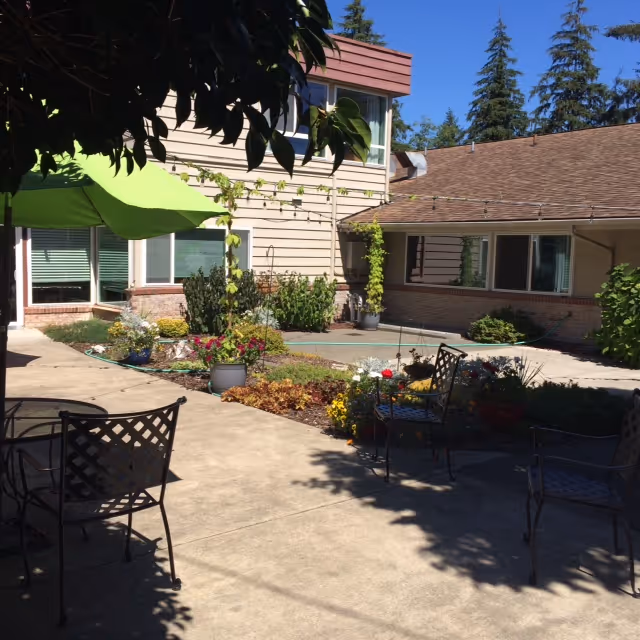 Outdoor courtyard area with metal chairs and tables, a green umbrella, various potted plants and flowers, surrounded by a two-story building with windows and trees in the background under a clear blue sky.