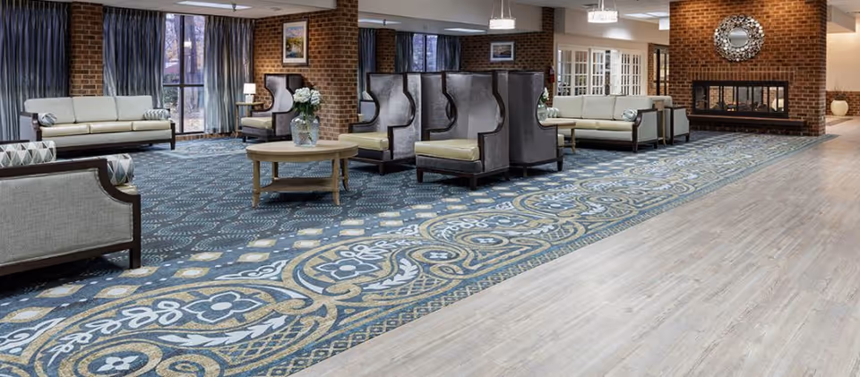 A spacious and well-lit living room area with patterned carpet and light wood flooring. The room features multiple seating options including sofas and high-backed armchairs arranged around a central round wooden table with a flower vase. Large windows with curtains allow natural light to enter, and a brick fireplace with a decorative mirror above it is visible on the right side.