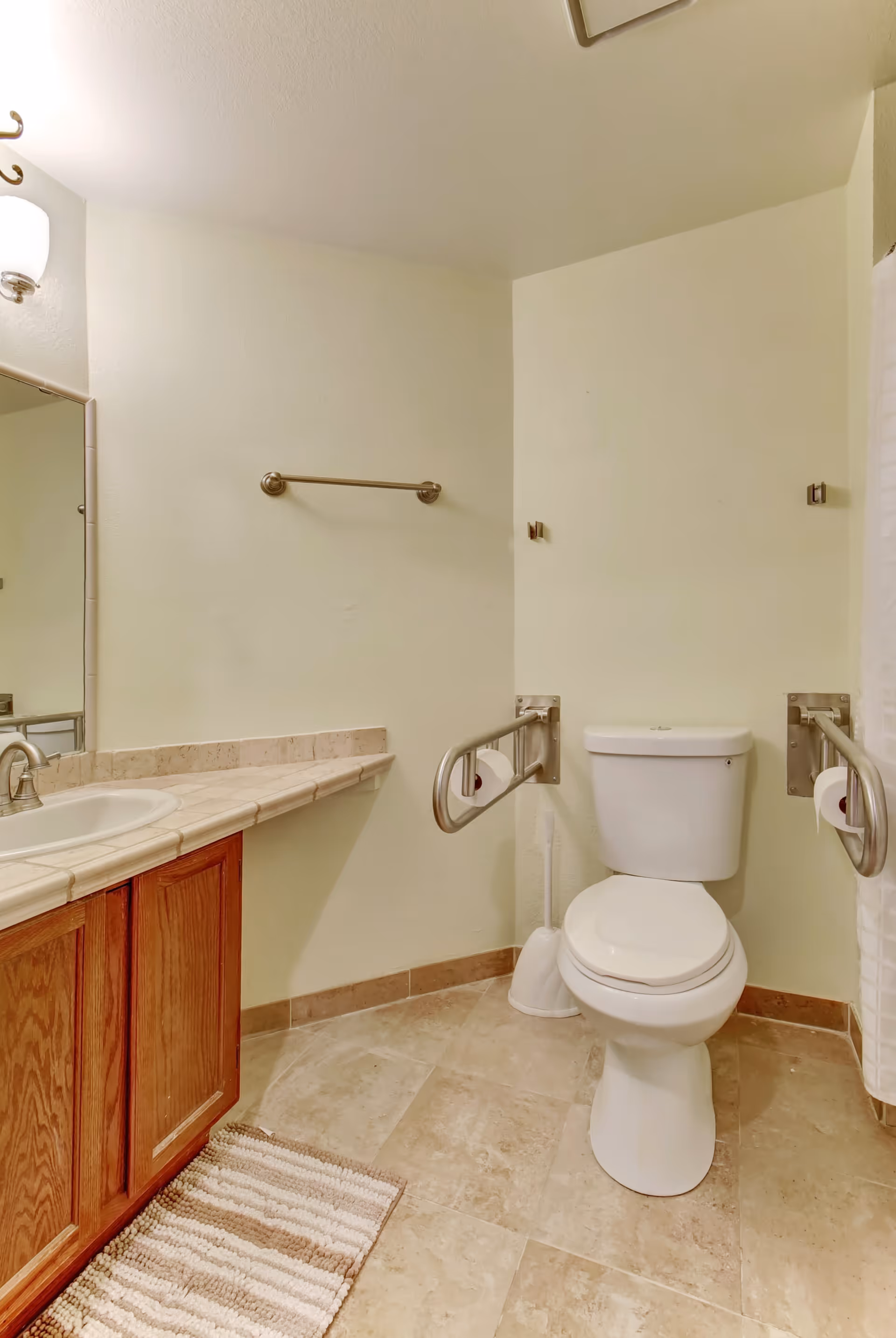 A clean bathroom with a white toilet equipped with metal grab bars on both sides for accessibility. There is a white toilet brush next to the toilet. To the left, there is a wooden vanity with a tiled countertop and a sink. Above the sink is a mirror and a wall-mounted light fixture. The floor is tiled and there is a striped bath mat in front of the vanity.